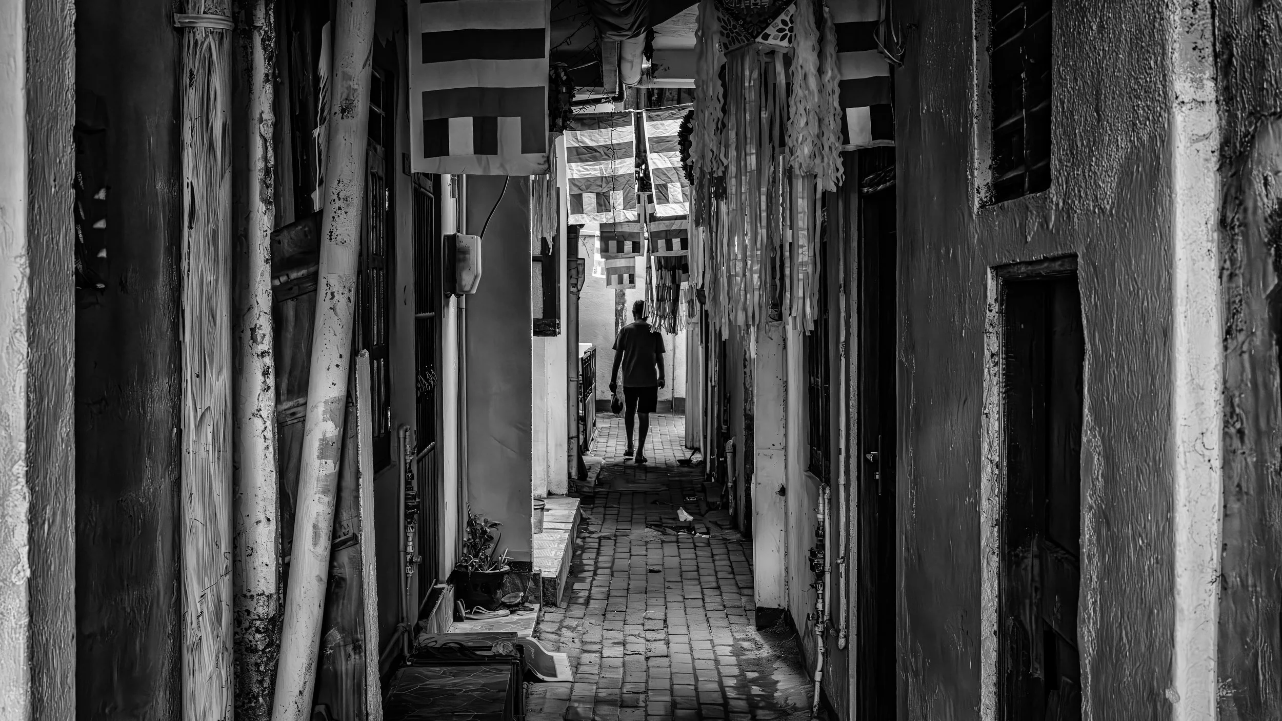 A narrow alleyway with textured walls on each side and hanging flags or banners overhead. A person is walking away from the camera down the cobblestone path.