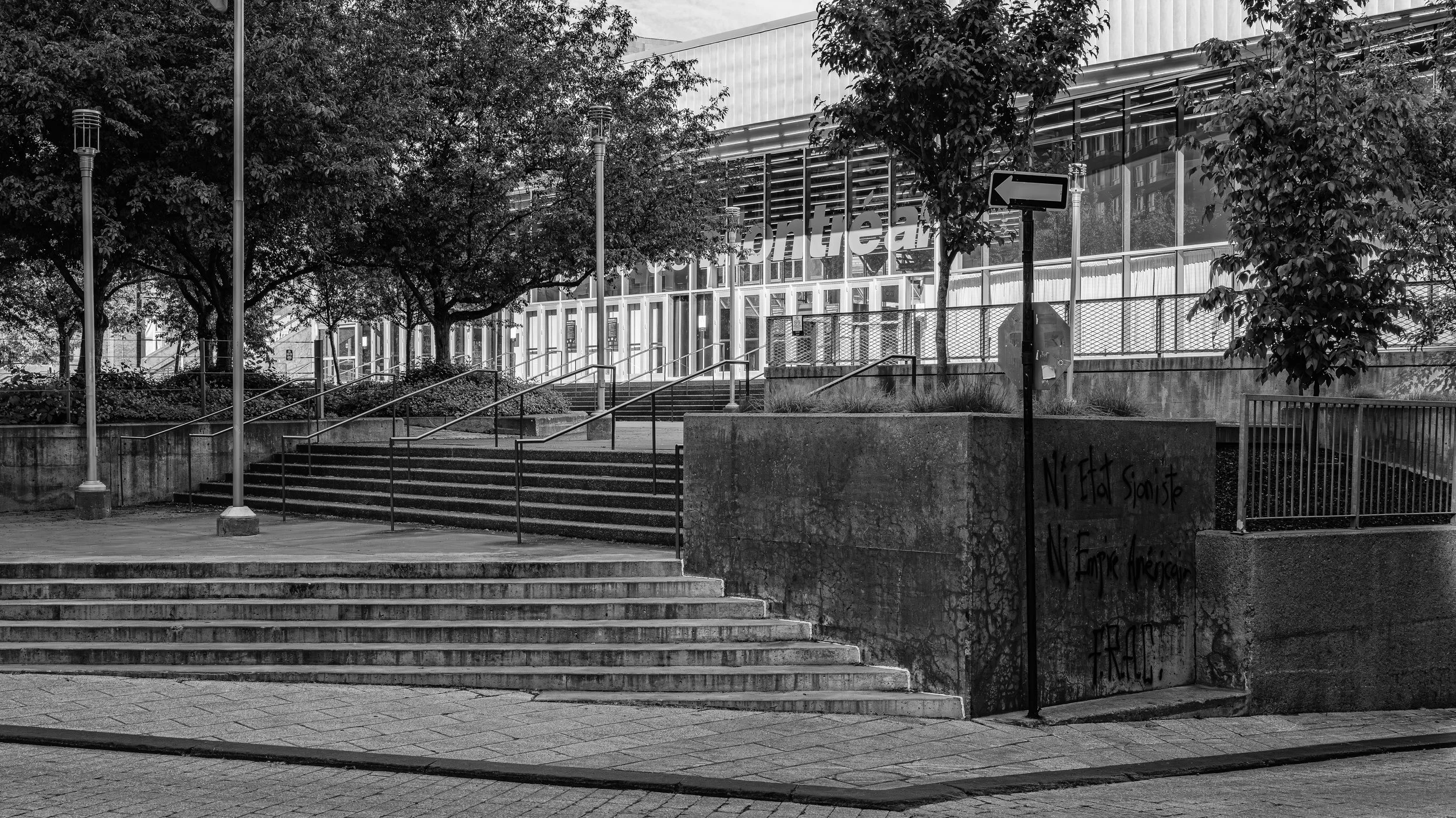 Black and white image of stairs leading up to a modern building with trees and street lamps in front. The building has large windows and a sign reading 'frontier'.