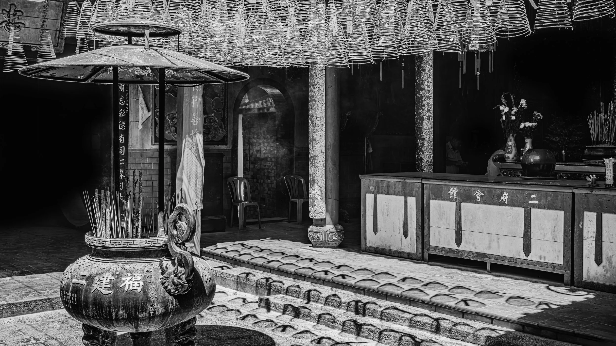 Black and white photo of a traditional Asian temple or shrine interior with decorative columns, hanging lanterns, vases with flowers, a stone incense burner with incense sticks, and a stone altar or counter with Chinese or Japanese characters.