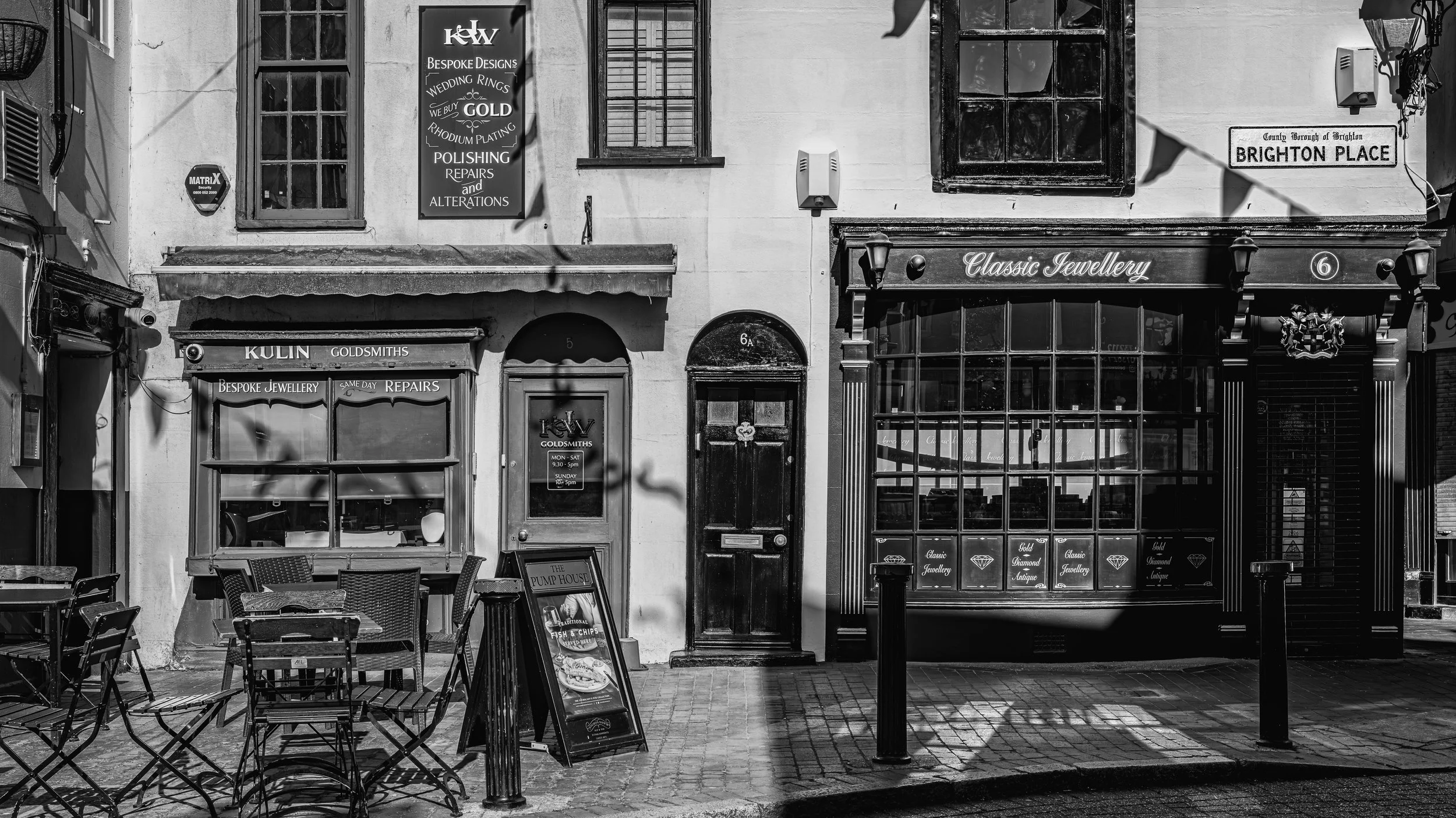 A black and white street scene showing storefronts and signs, including a jewelry shop, a repair shop, and a pub with outdoor seating and chairs.