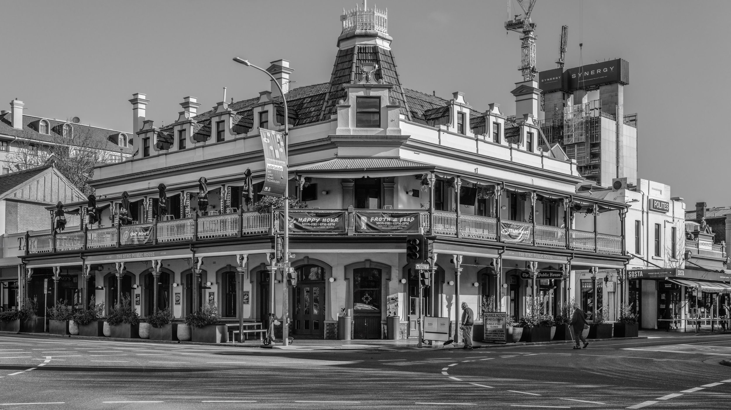 Black and white photo of a corner building with balcony, decorative roof, and advertisements for events, with street and pedestrians in the foreground. Adelaide, Australia.