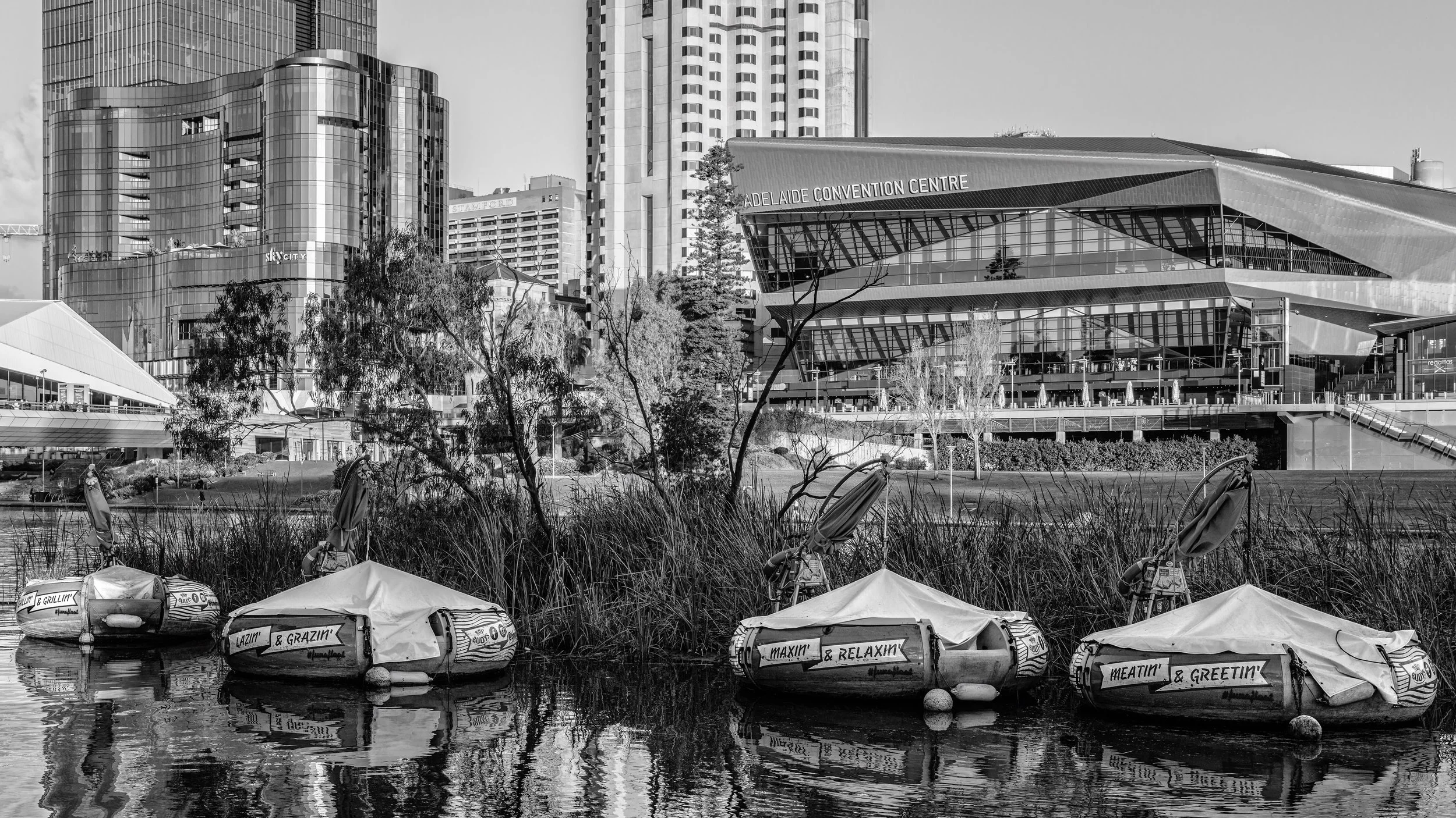 Boats docked on a calm river with a cityscape in the background, including modern high-rise buildings and the Adelaide Convention Centre, in black and white. Adelaide, Australia.