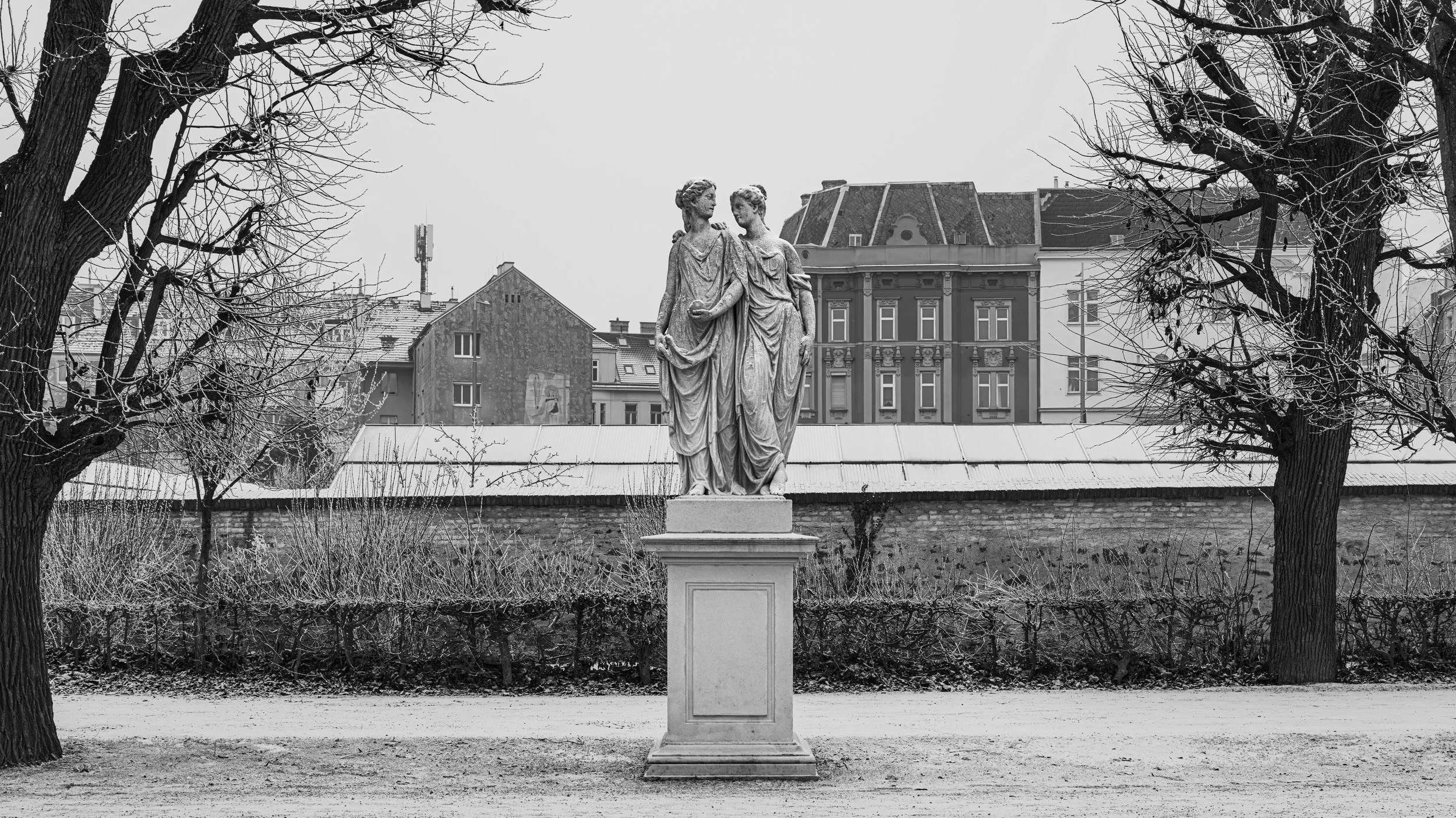 Black and white photo of a stone statue of two women in a park, flanked by leafless trees with buildings in the background.