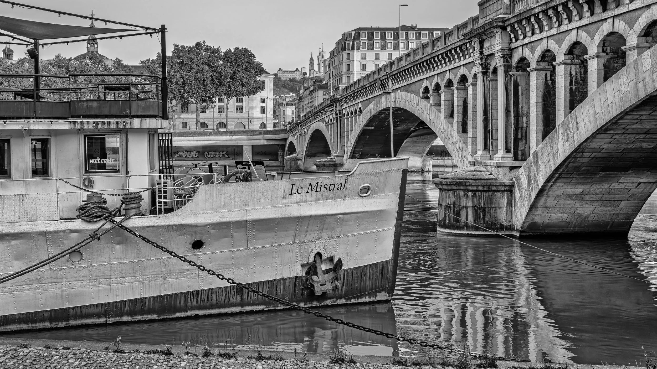 Black-and-white photo of a docked boat named "Le Mistral" in a harbor with an arched stone bridge in the background, and buildings on a hillside behind the bridge. Lyon, France.