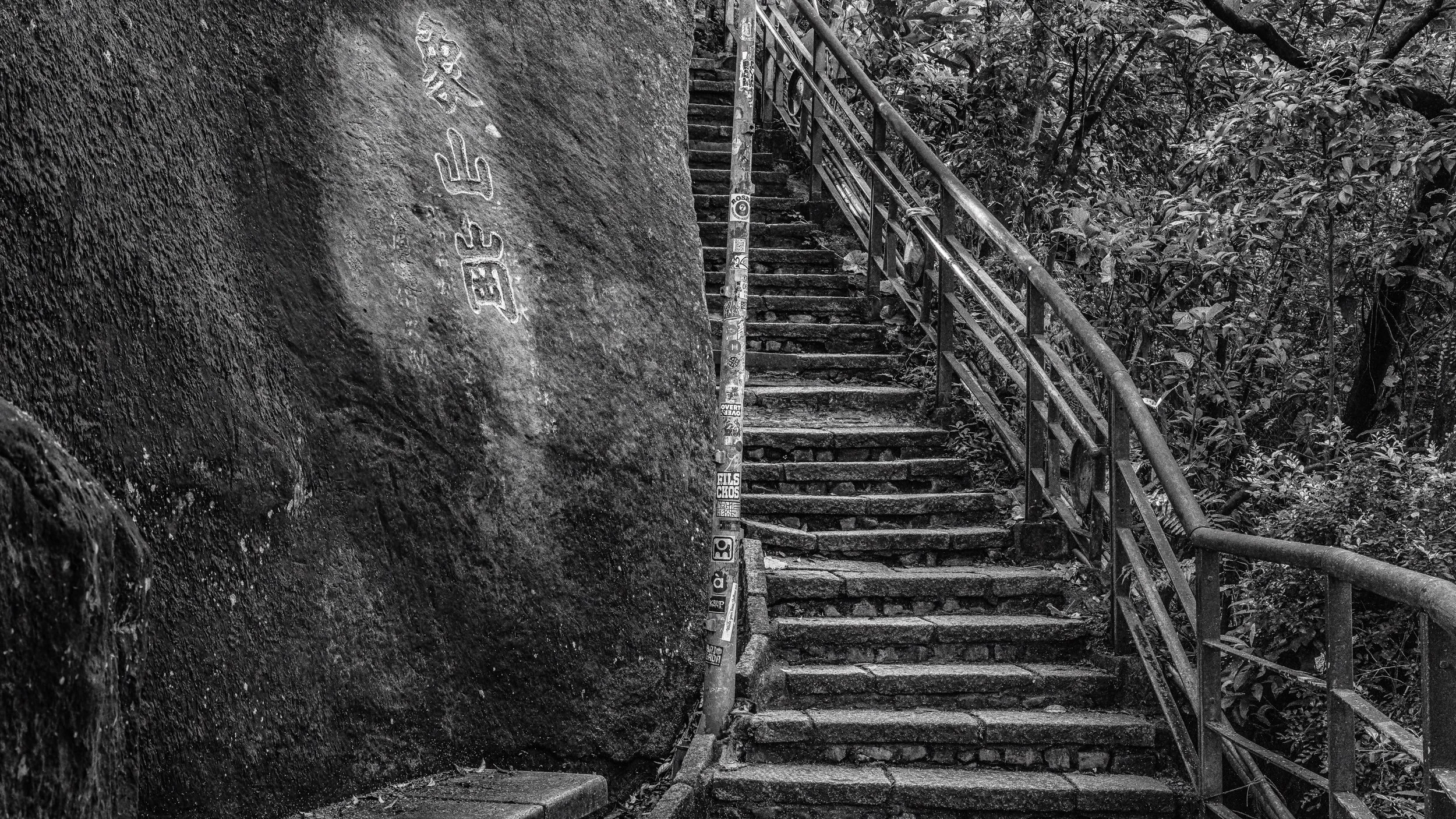 A black and white photo of a staircase made of concrete steps with metal railings, ascending alongside a large rock face covered in graffiti and stickers in a wooded area.