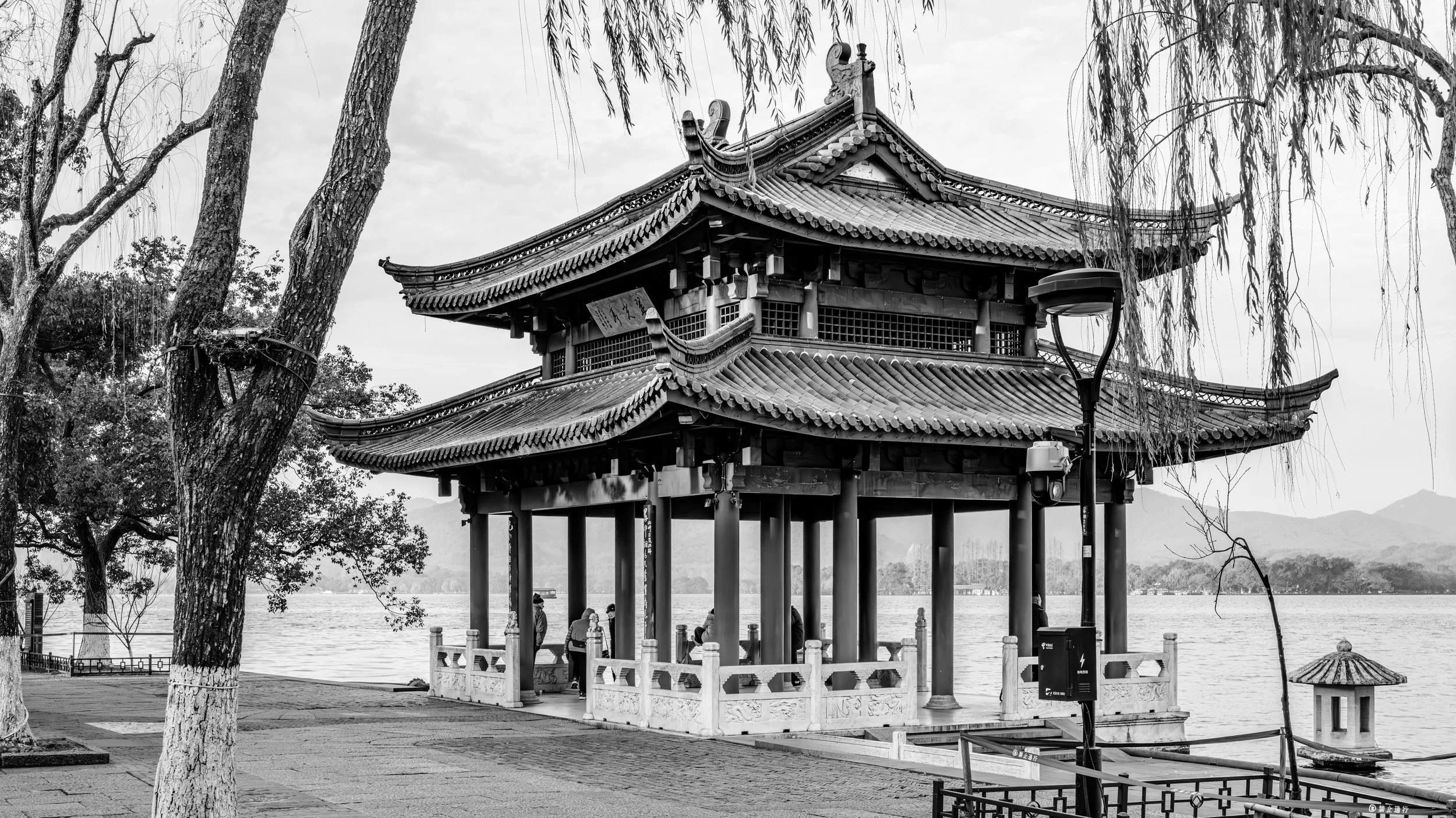 Traditional Asian pavilion by a lake, with trees and mountains in the background, in black and white.