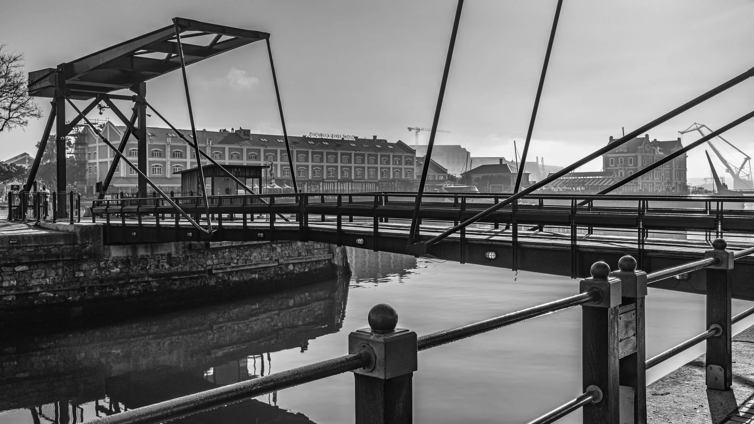 Black and white photo of a waterfront scene with a bridge, historic buildings, and cranes in the background. Cape Town, South Africa.