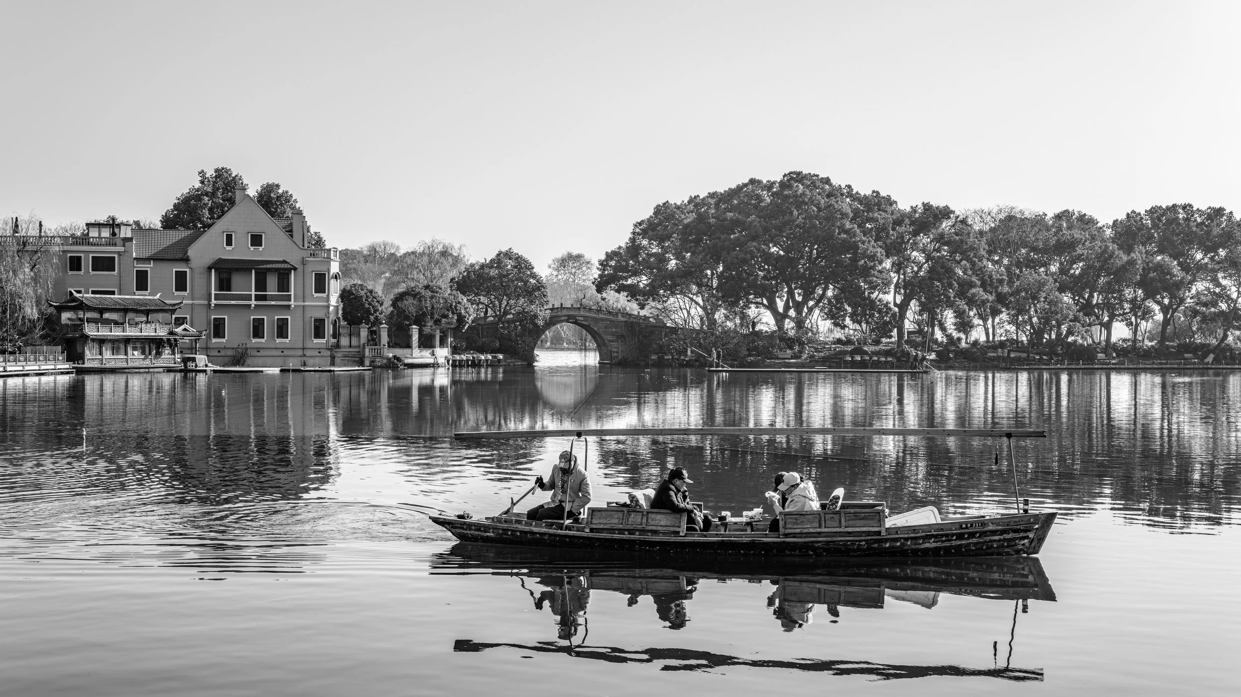 People fishing from a small boat on a calm river, with traditional architecture and a bridge in the background, and trees along the riverbank.