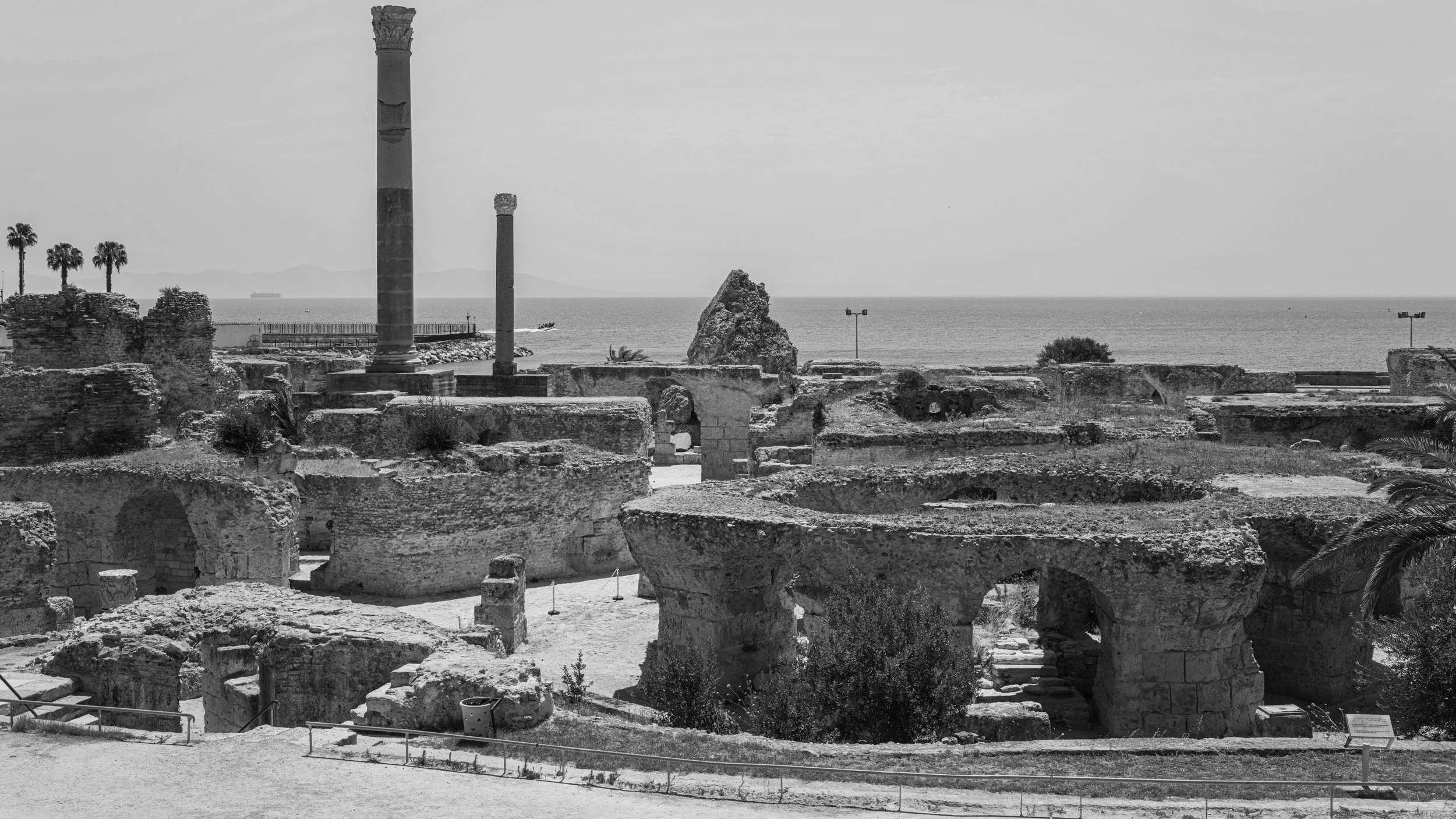 Ancient ruins with stone columns and walls overlooking the ocean, with a large rock formation in the sea and distant mountains in the background.