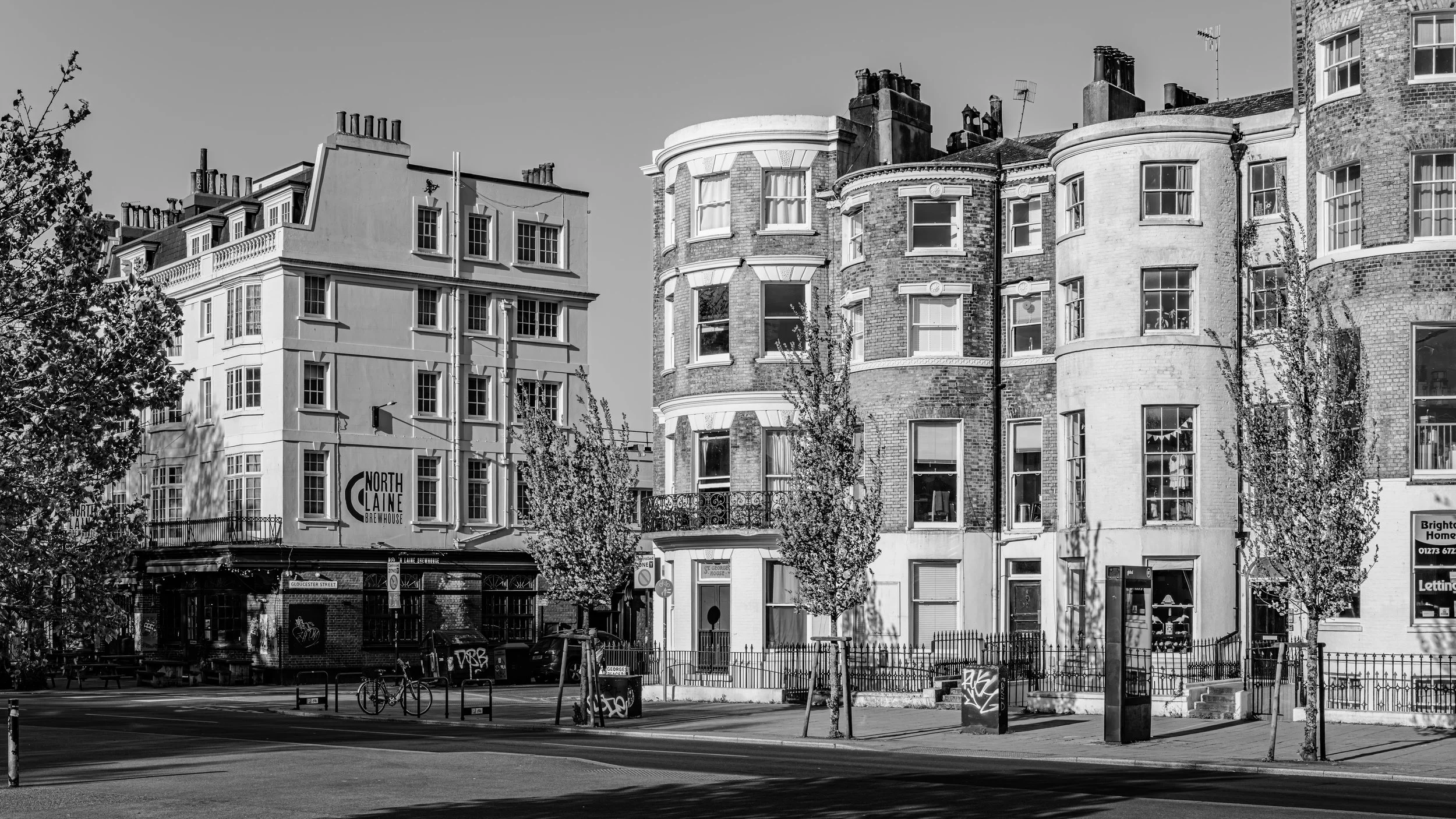 Black and white photo of historic brick and stucco buildings on a city street, with trees, bicycles, and street signs in the foreground.