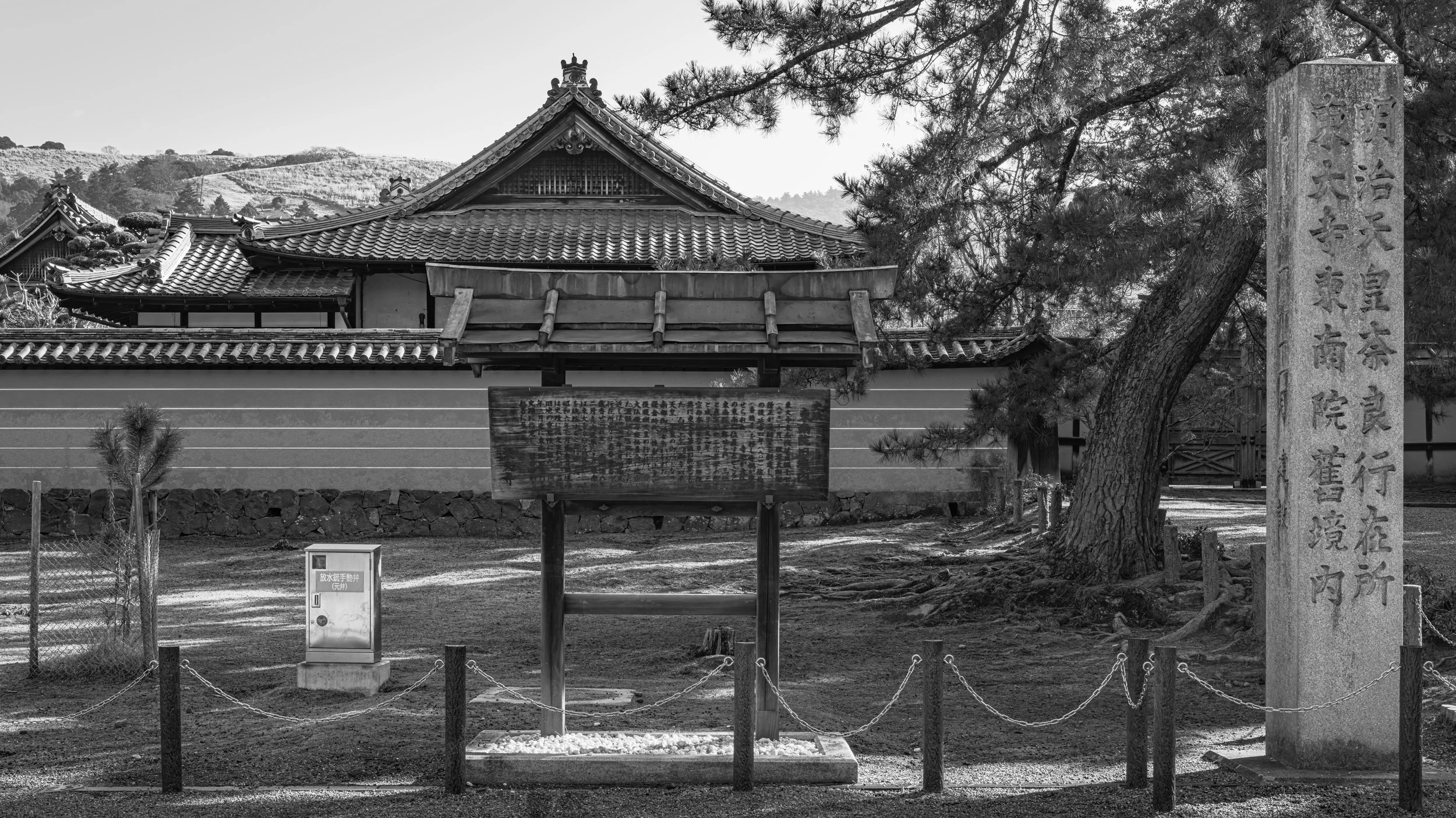 A traditional Japanese temple with tiled roofs, surrounded by trees and a mountain landscape in the background. There is a tall stone monument with Japanese inscriptions on the right, and a board with text in front of the temple grounds. The scene is