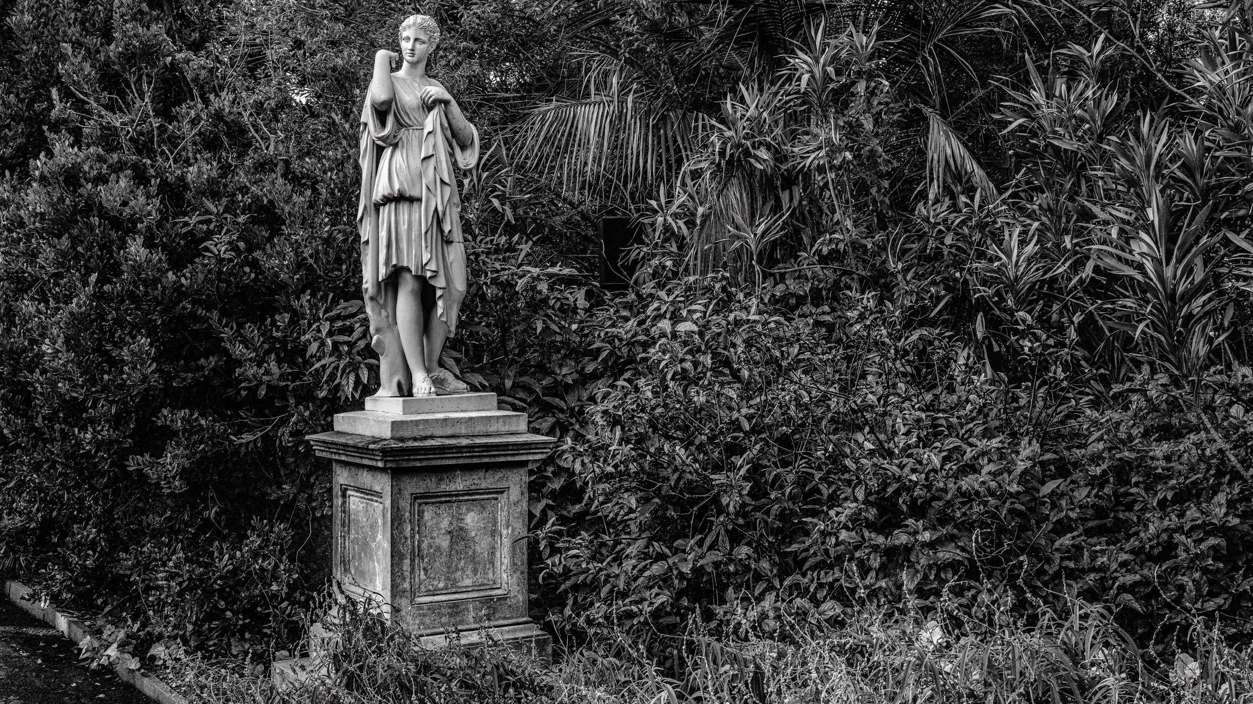 A stone statue of a woman in a flowing dress stands atop a pedestal in a lush garden with dense foliage behind it. Adelaide, Australia.