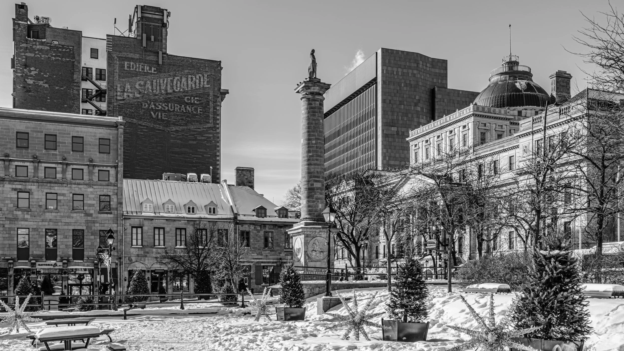 Black and white photo of a cityscape with historic and modern buildings, a statue on a column, leafless trees, and snow-covered ground in a park.