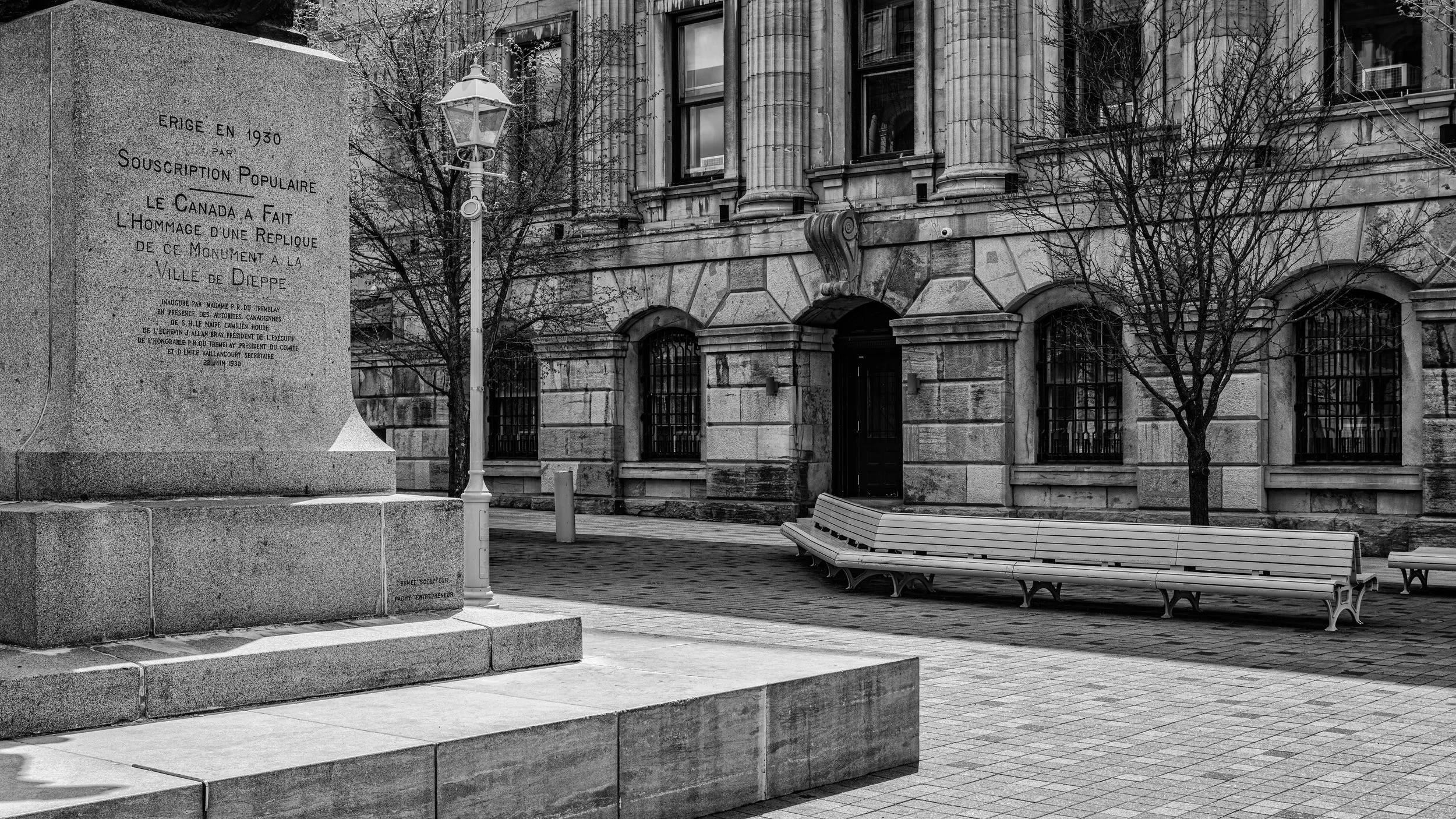 A black and white photo of a city street scene featuring a large stone monument with an inscription, several leafless trees, a curved streetlamp, a bench, and a historic building with columns and arched windows.