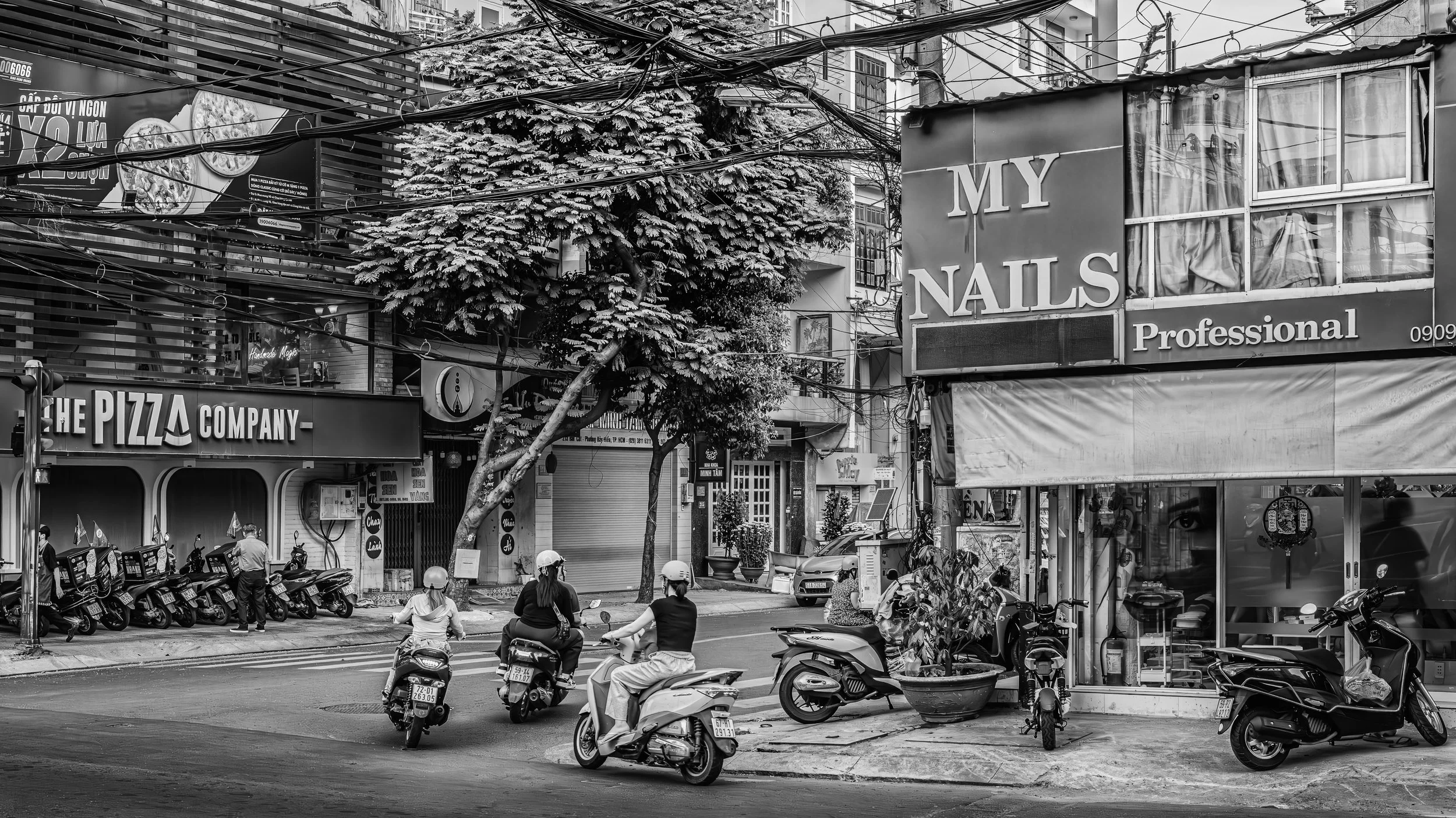 A bustling city street scene in black and white with multiple motorcycles and scooters on the road, pedestrians walking, and storefronts including a nail salon and pizza shop. There are trees and a tangle of overhead electrical wires in the scene.