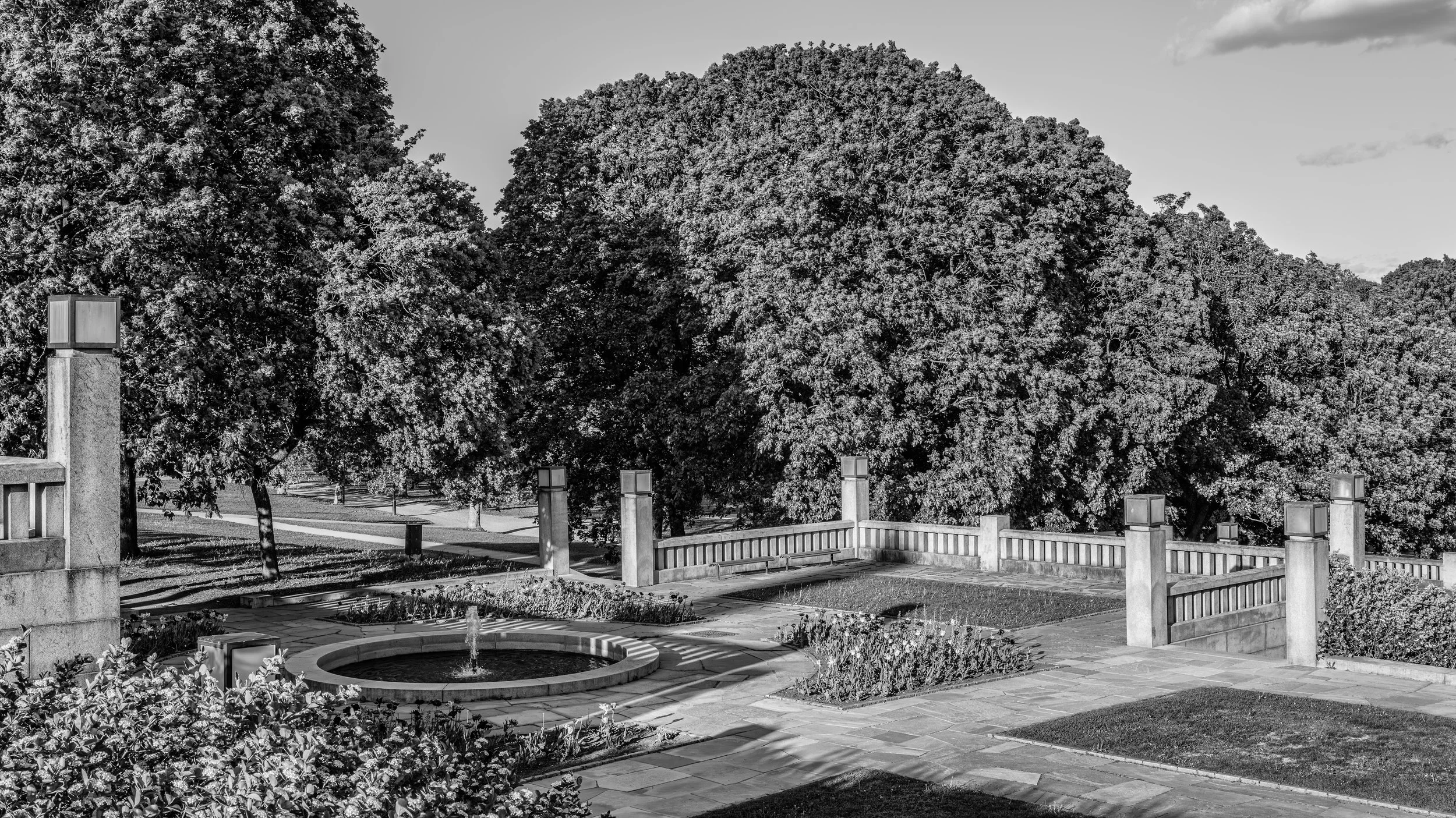A park scene with large leafy trees, a fountain in a circular flower bed, and a paved area with lamp posts and a wooden fence in black and white.