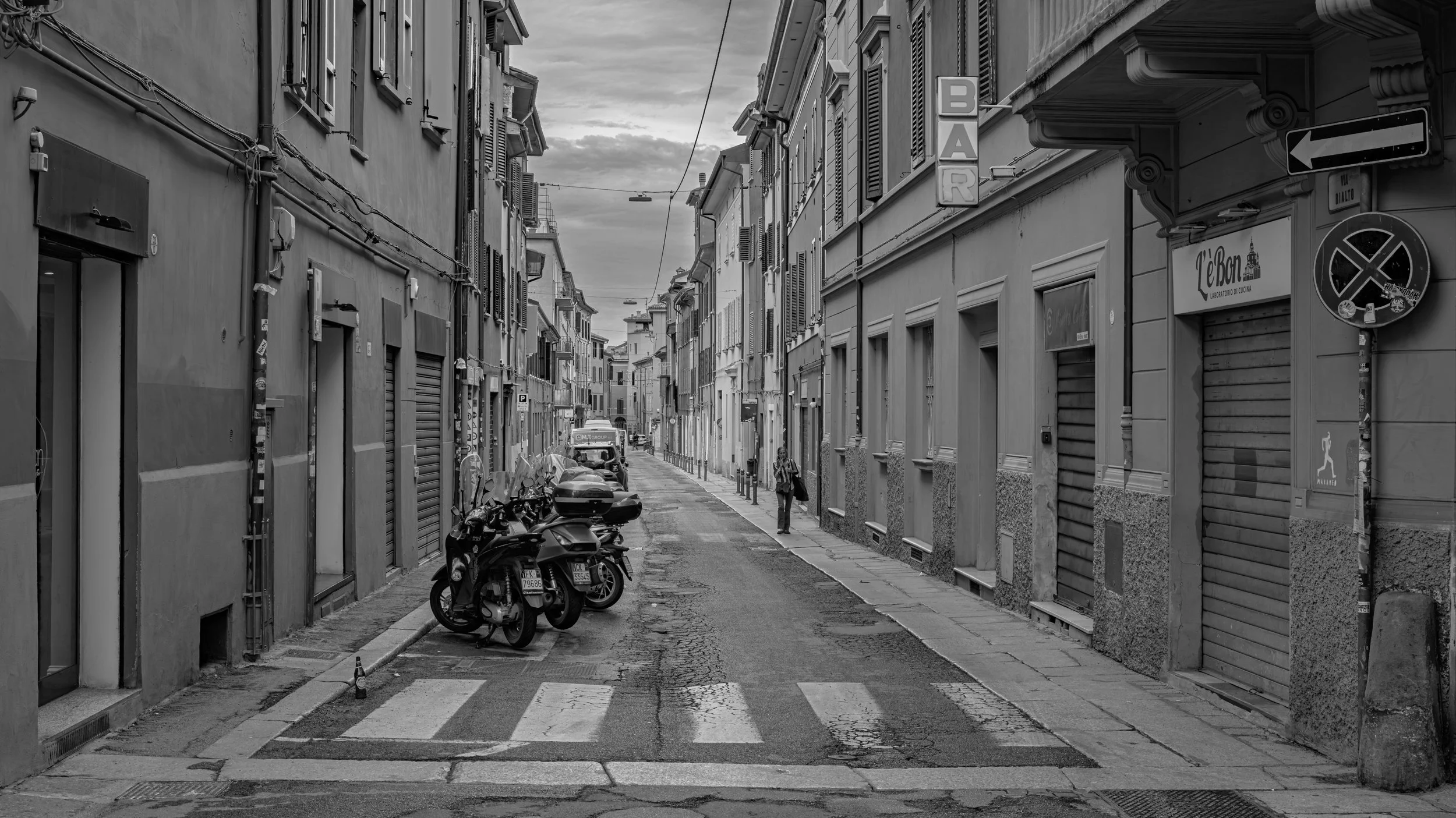 A narrow empty street in a European city, with parked motorcycles and a woman walking in the distance, surrounded by tall buildings with shuttered windows.