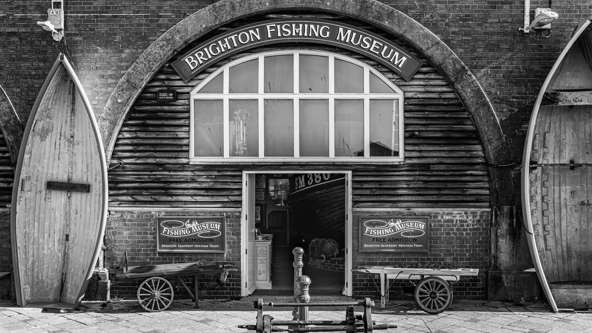 Black and white photo of the Brighton Fishing Museum with an arched entrance, two boats on either side, and signs indicating free admission and the Brighton Seafront Heritage Trust.