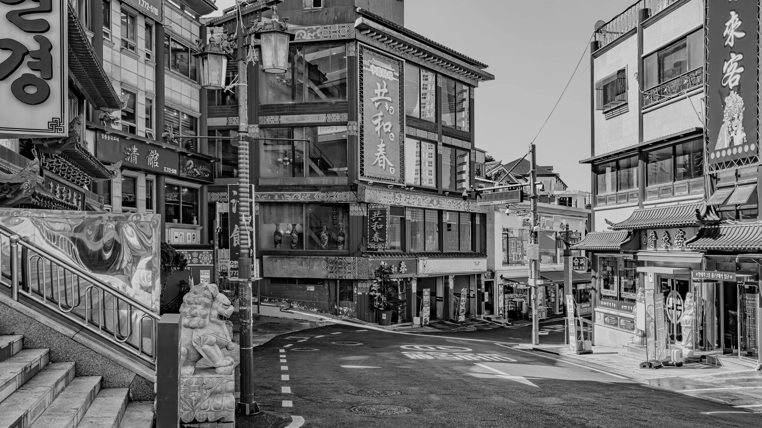 Black and white photo of a busy city street with multiple buildings displaying Asian signage. There is a stone lion statue near stairs at the foreground, and several lanterns hanging from poles.