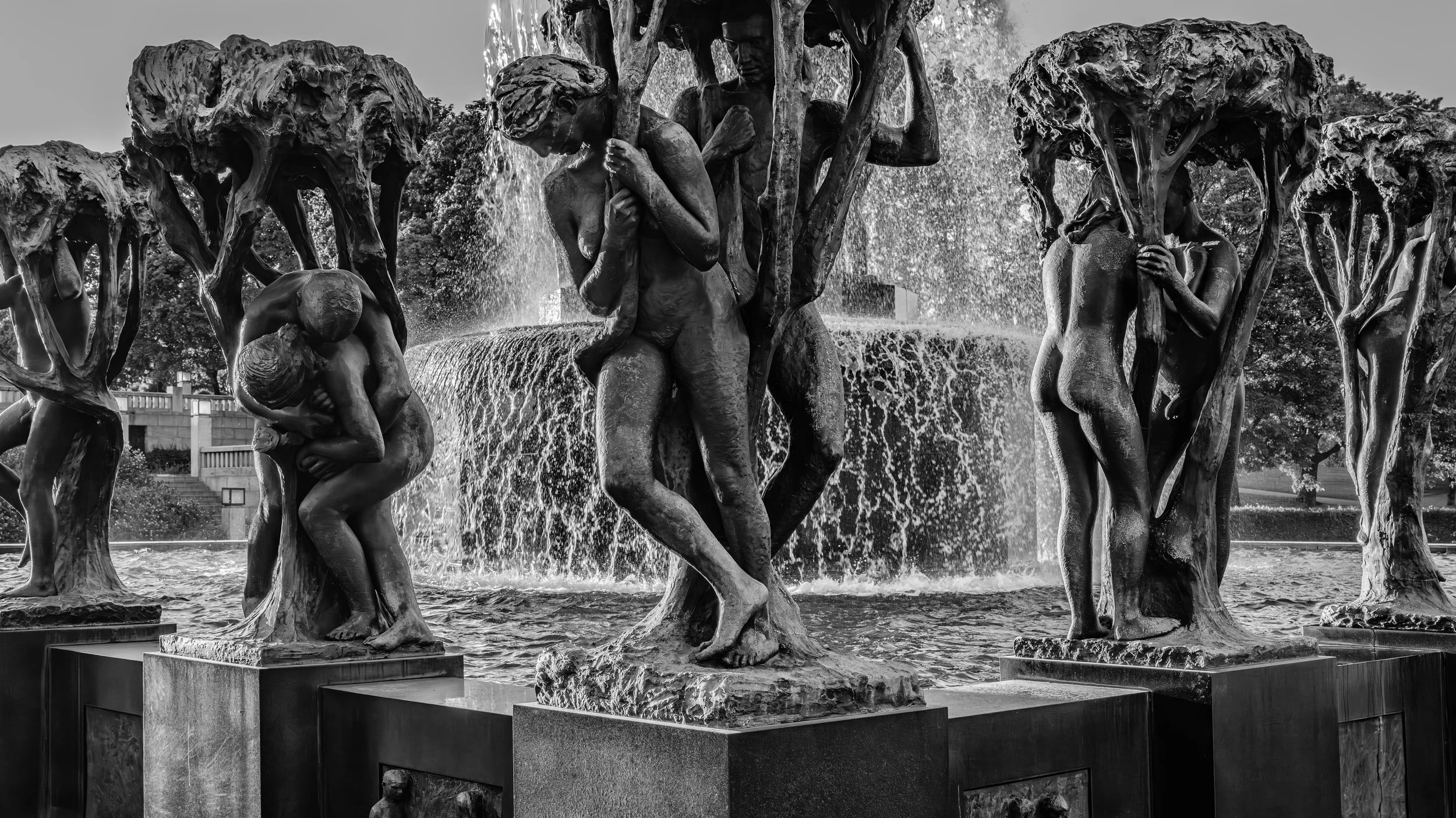 A black and white photo of a fountain with multiple bronze statues of human figures, some nudely, engaging in various poses and interactions, with water cascading around them.