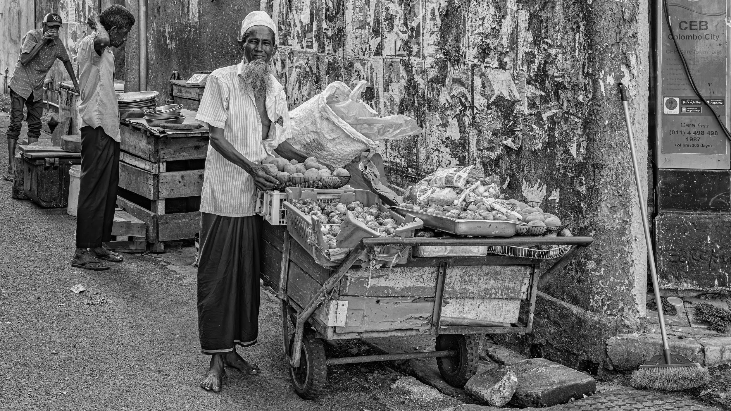 Three men selling fruits at a street market. One man stands behind a cart filled with apples and onions, smiling at the camera. Two other men are to the left, one on a stool and one leaning over a stand with plates. A weathered wall and a public phon
