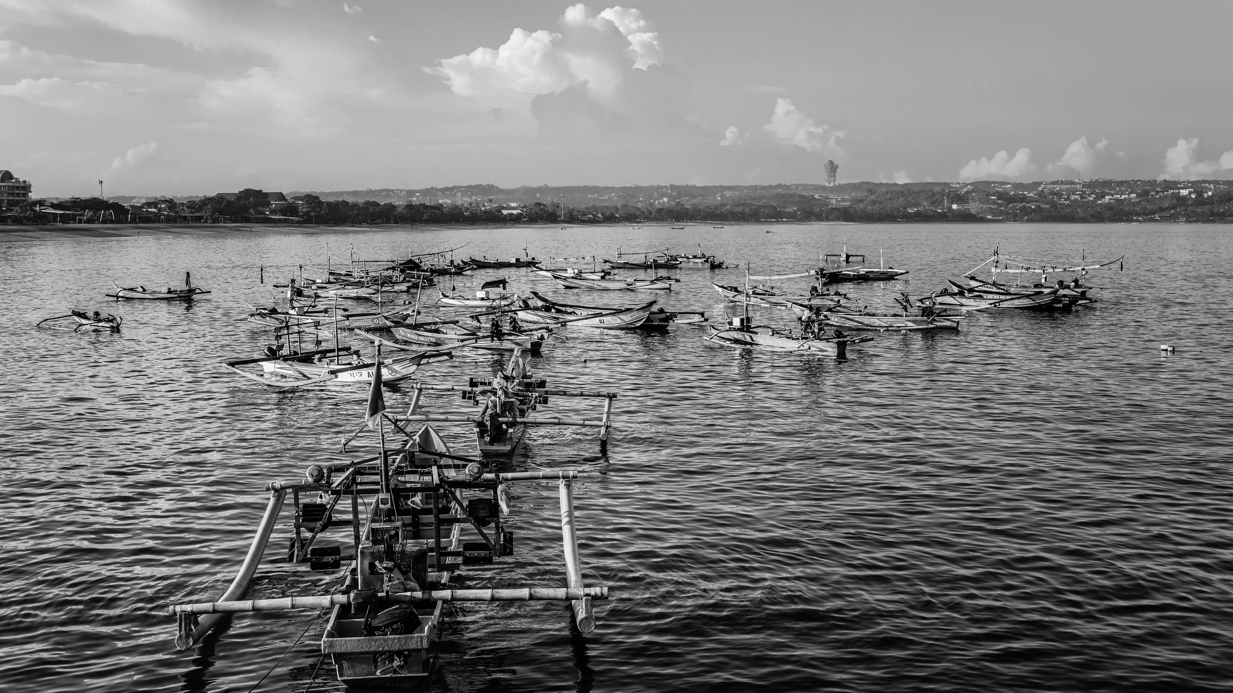 Black and white photo of boats docked on a calm body of water with a distant shoreline and scattered clouds in the sky.
