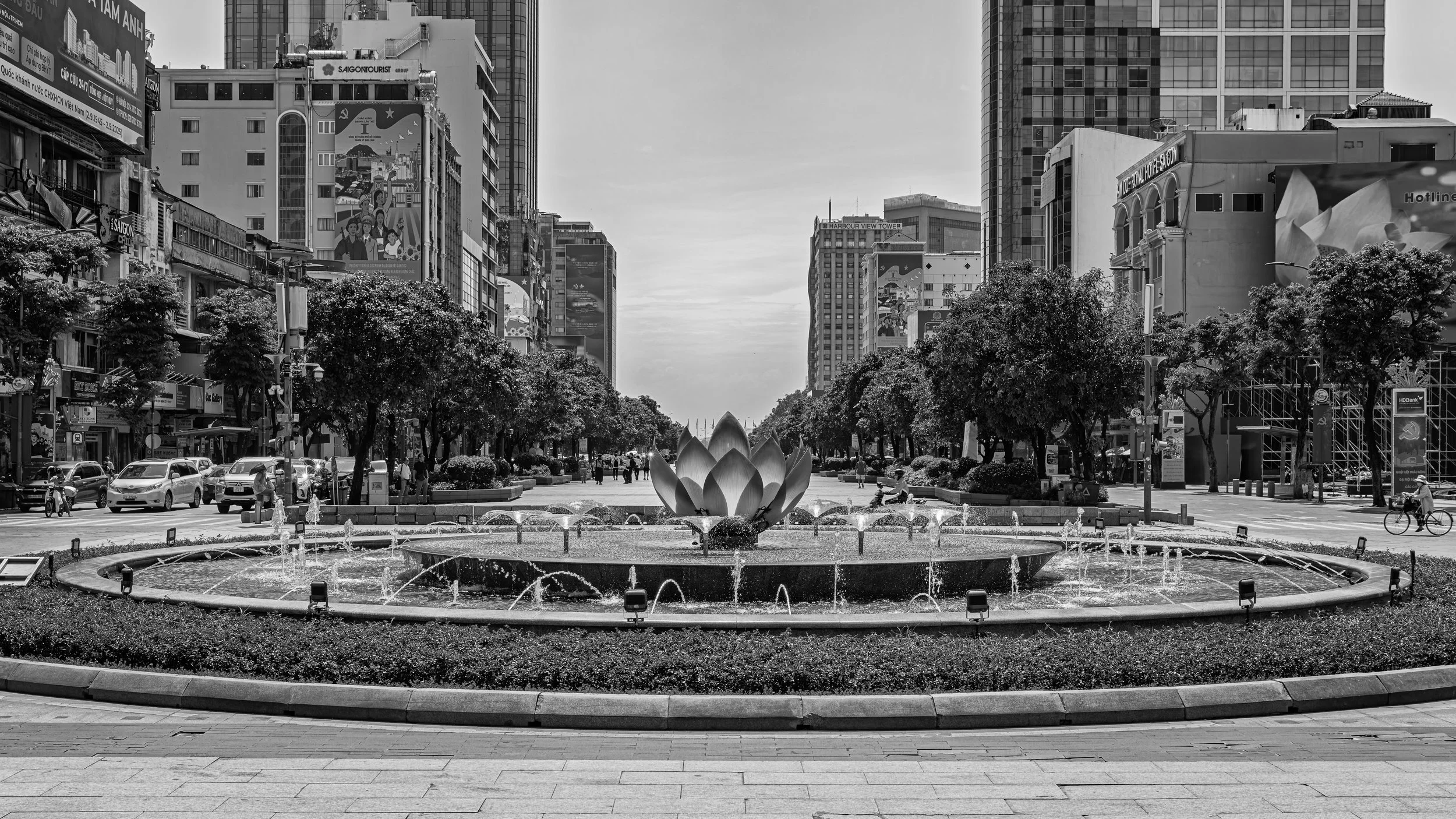 Black and white photograph of a city street with a fountain in the foreground, surrounded by trees, buildings, and people walking and biking.
