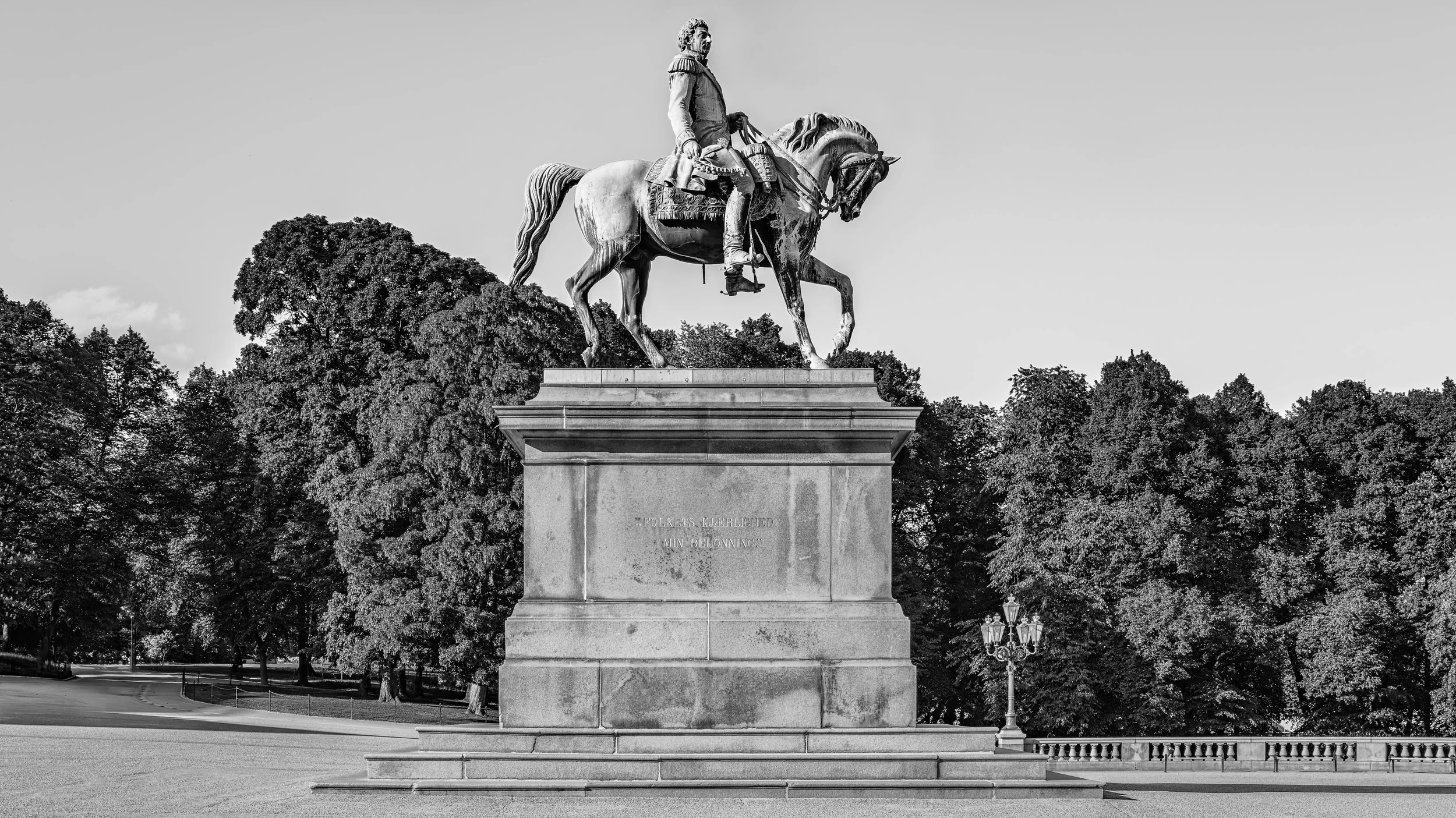 A black and white statue of a man in military uniform on a horse, standing on a large stone pedestal surrounded by trees.