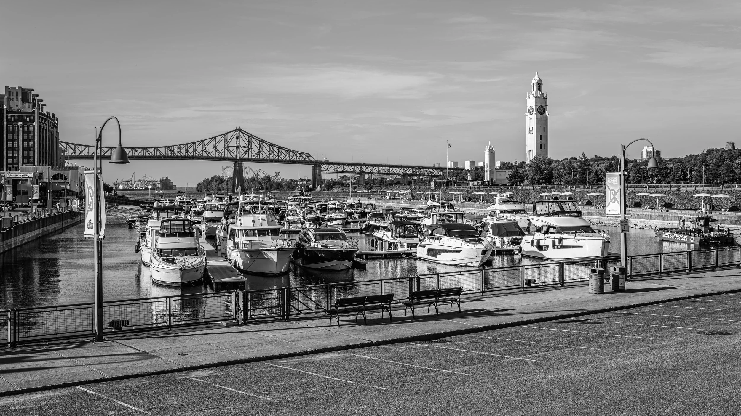 A harbor with boats docked, a bridge in the background, and a tall clock tower, all in black and white.