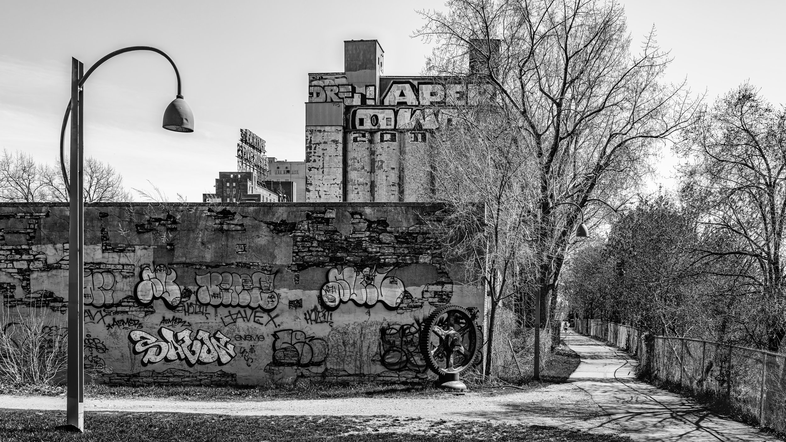 Black and white photo of an old, graffiti-covered brick wall with graffiti art, a large gear, and a partially visible building with silo-like structures in the background. There are leafless trees and a sidewalk alongside the wall, with a tall street