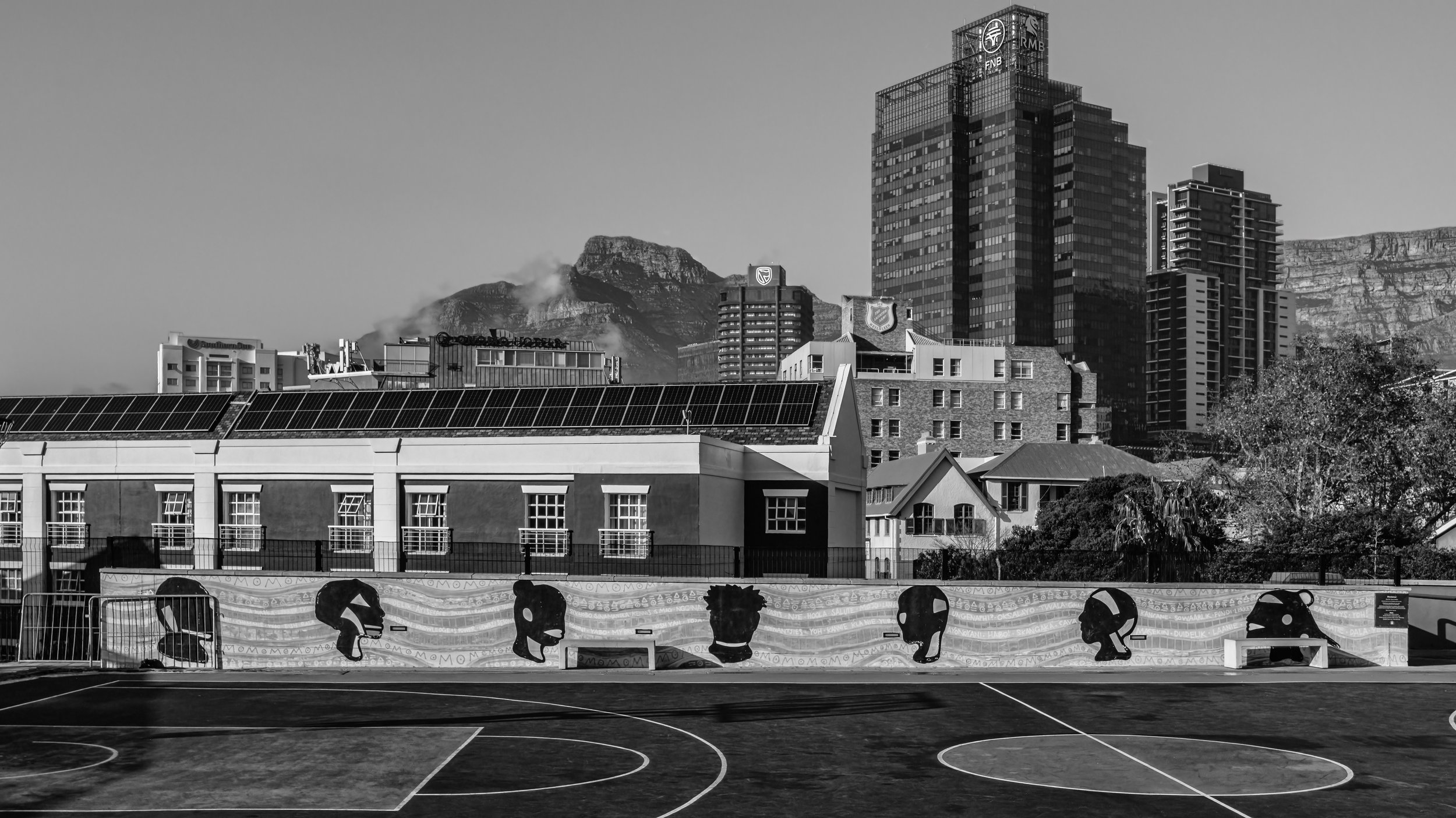 Black and white photo of a city skyline with tall modern buildings, a mountain in the background, and a fenced outdoor sports court or playground in the foreground. Cape Town, South Africa.