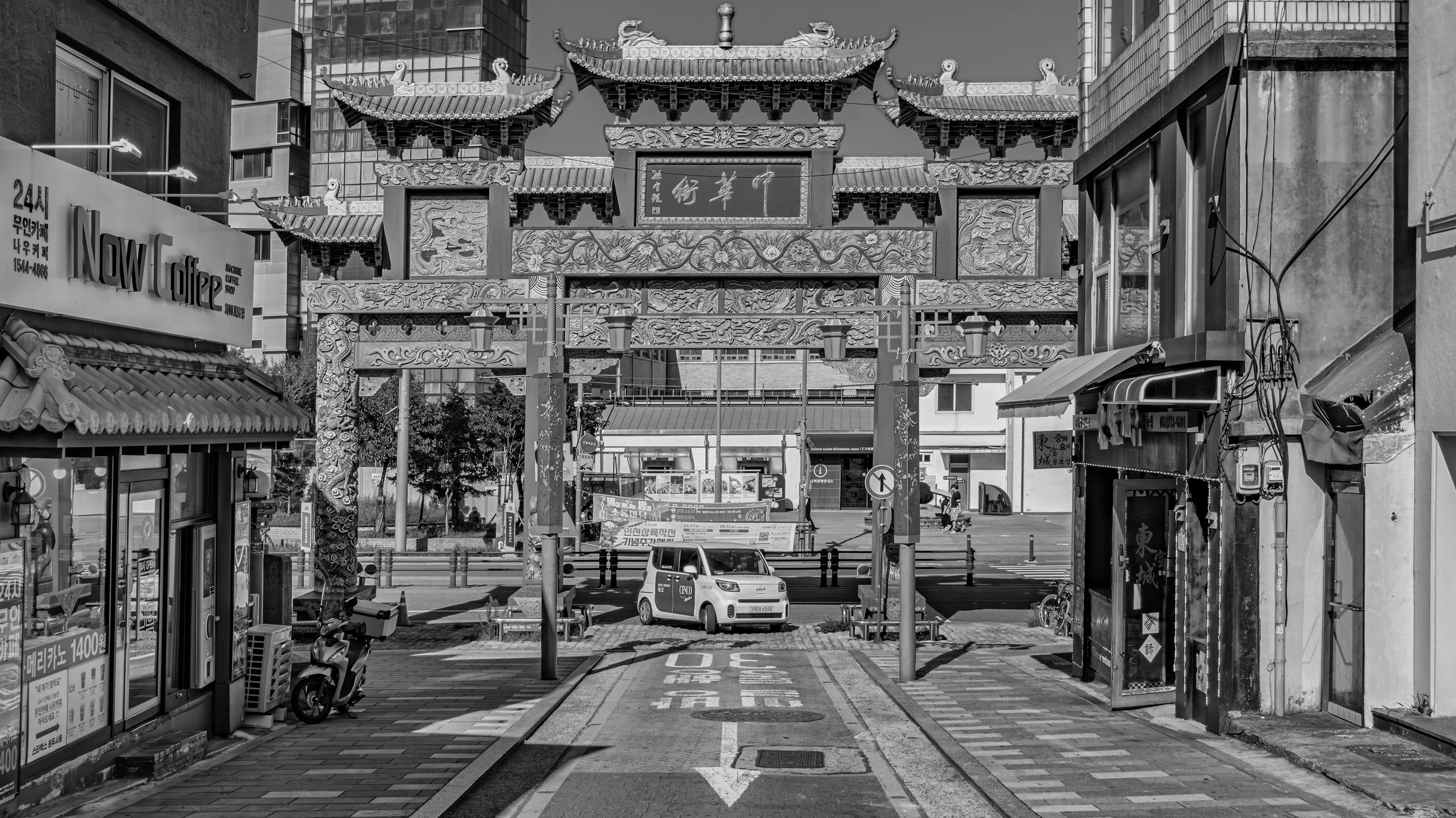 A black and white photo of a city street scene with traditional Chinese architectural gate and modern buildings. There are small vehicles, a scooter, and various signs visible.