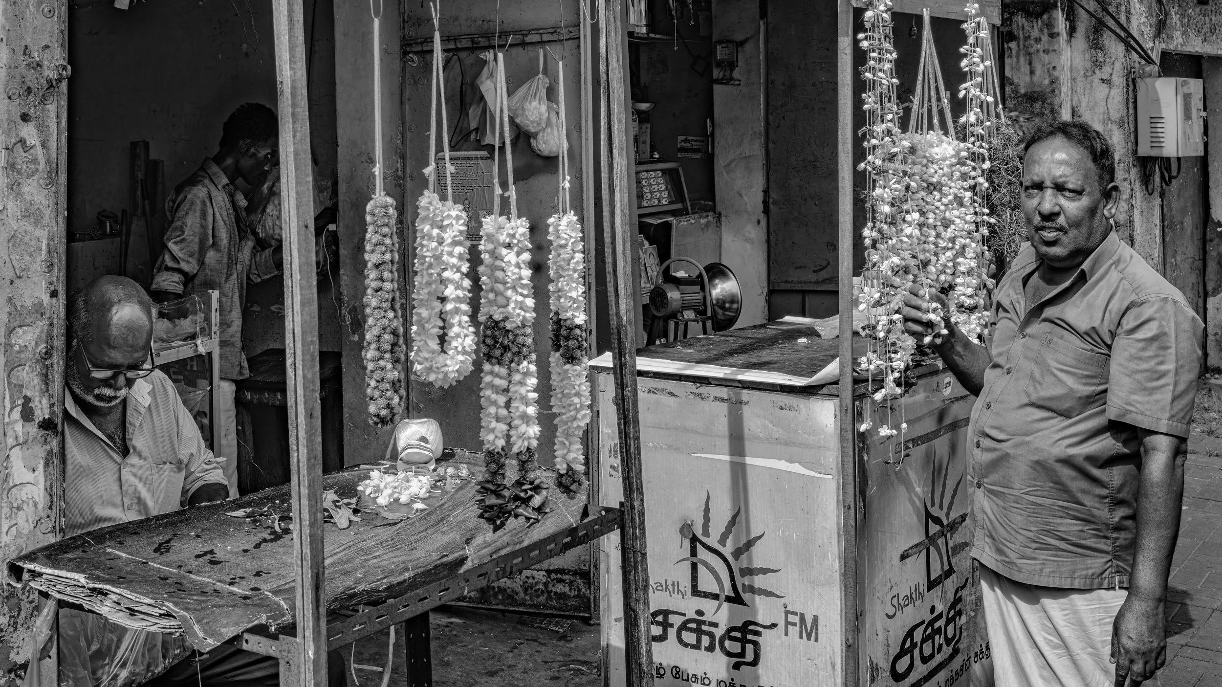 A street scene in black and white showing three men at a small flower stall. One man is sitting on the left, wearing glasses and a striped shirt, looking downward. Another man is standing on the right, holding and examining a floral garland, wearing 