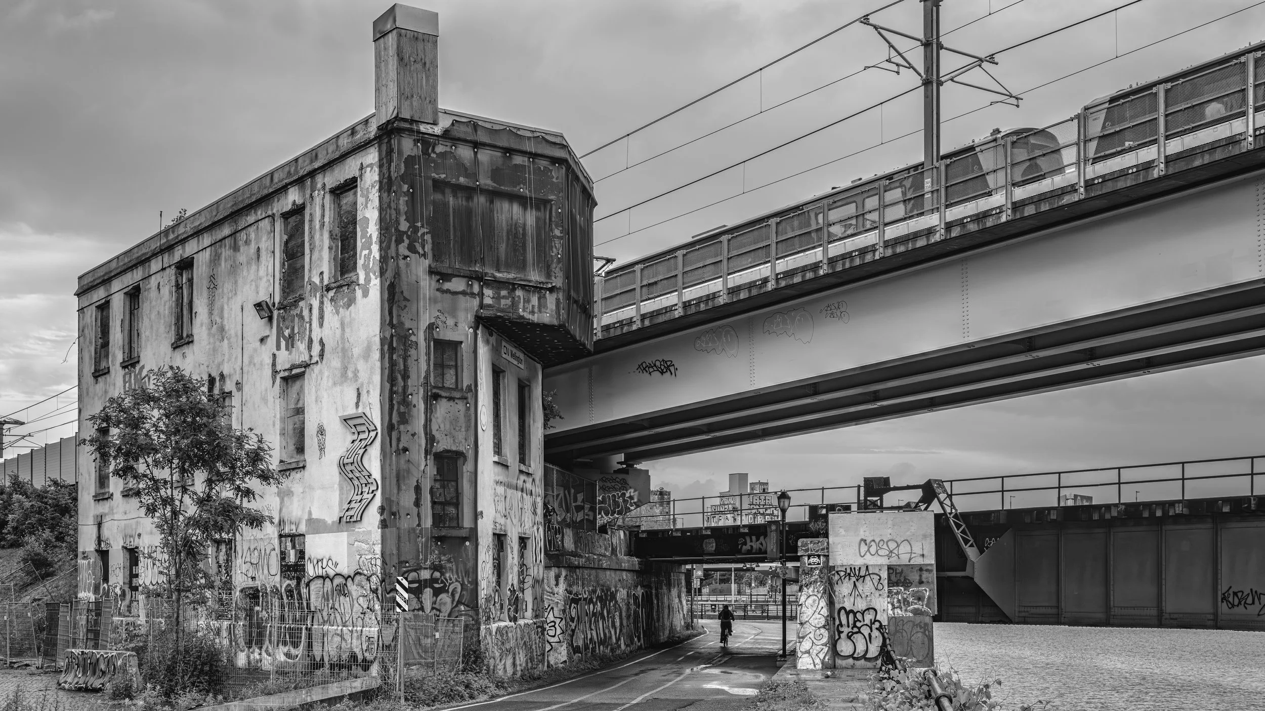 Black and white photo of an urban scene with an old, weathered building covered in graffiti, an overpass bridge above, and a cyclist riding through the area. Montreal, Canada.