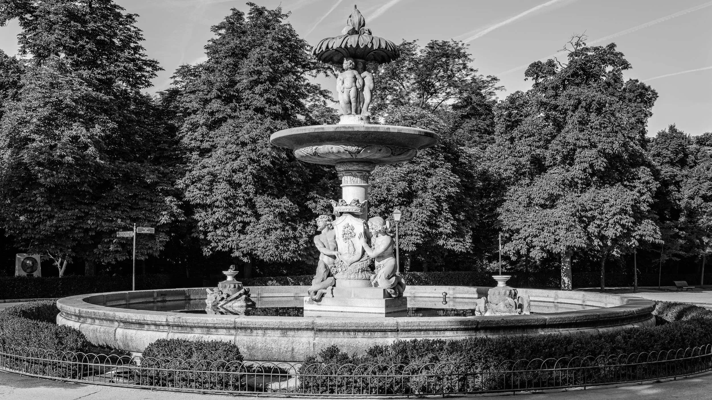A black and white photograph of an ornate multi-tiered fountain with sculptures, surrounded by trees and lampposts.