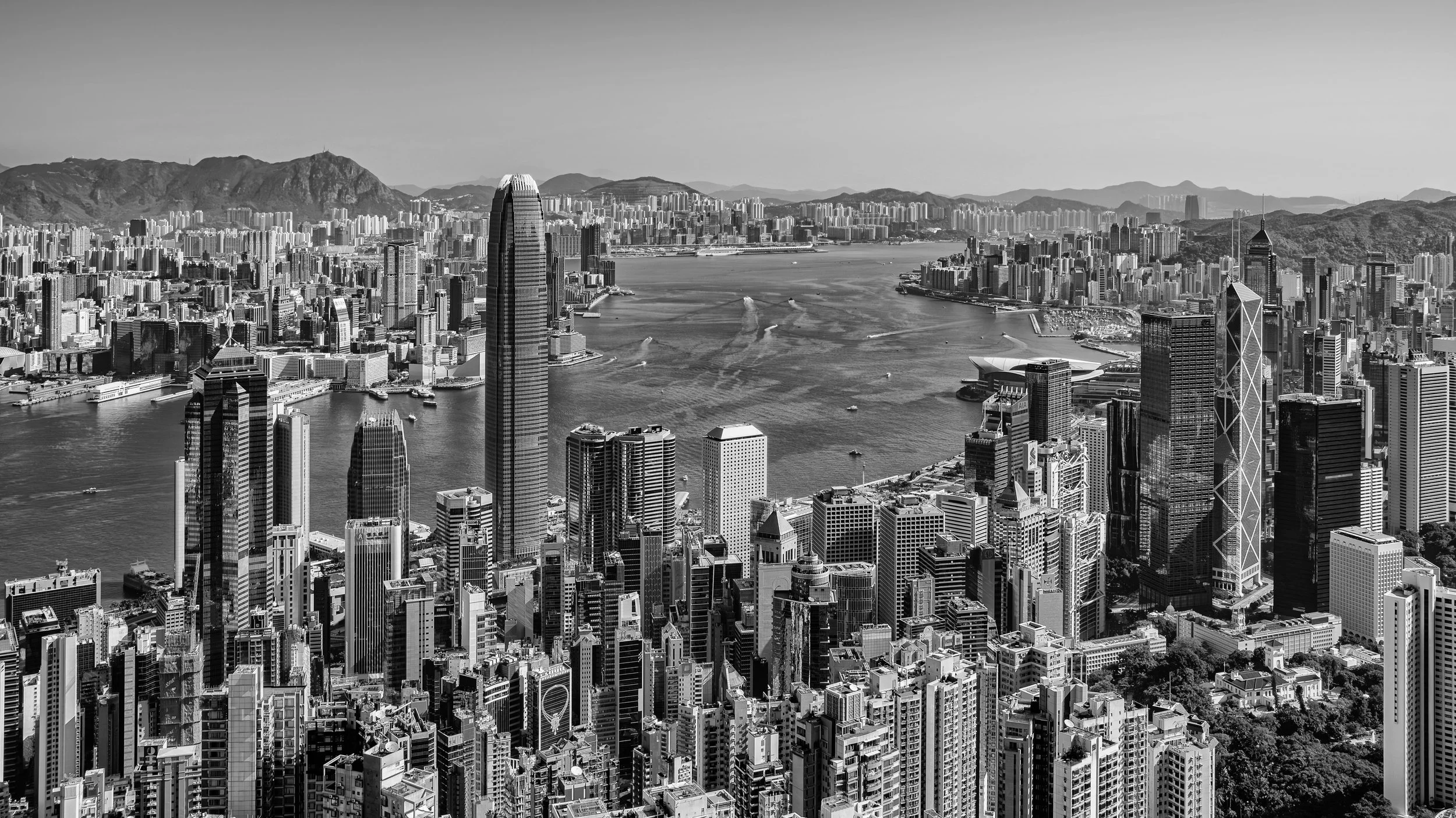 Black and white aerial view of a city skyline with tall skyscrapers, a river with boats, and mountains in the background.
