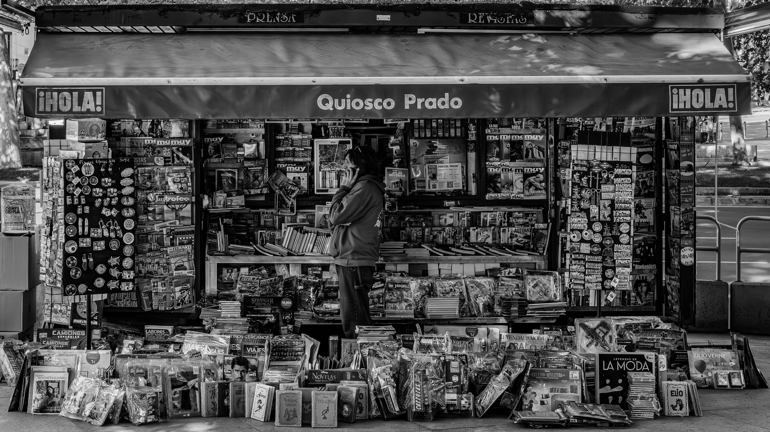 A street kiosk selling books, magazines, and stationery items. The kiosk has signs reading "¡HOLA!" and "Quesco Prado". A person is browsing at the stall, talking on a mobile phone.