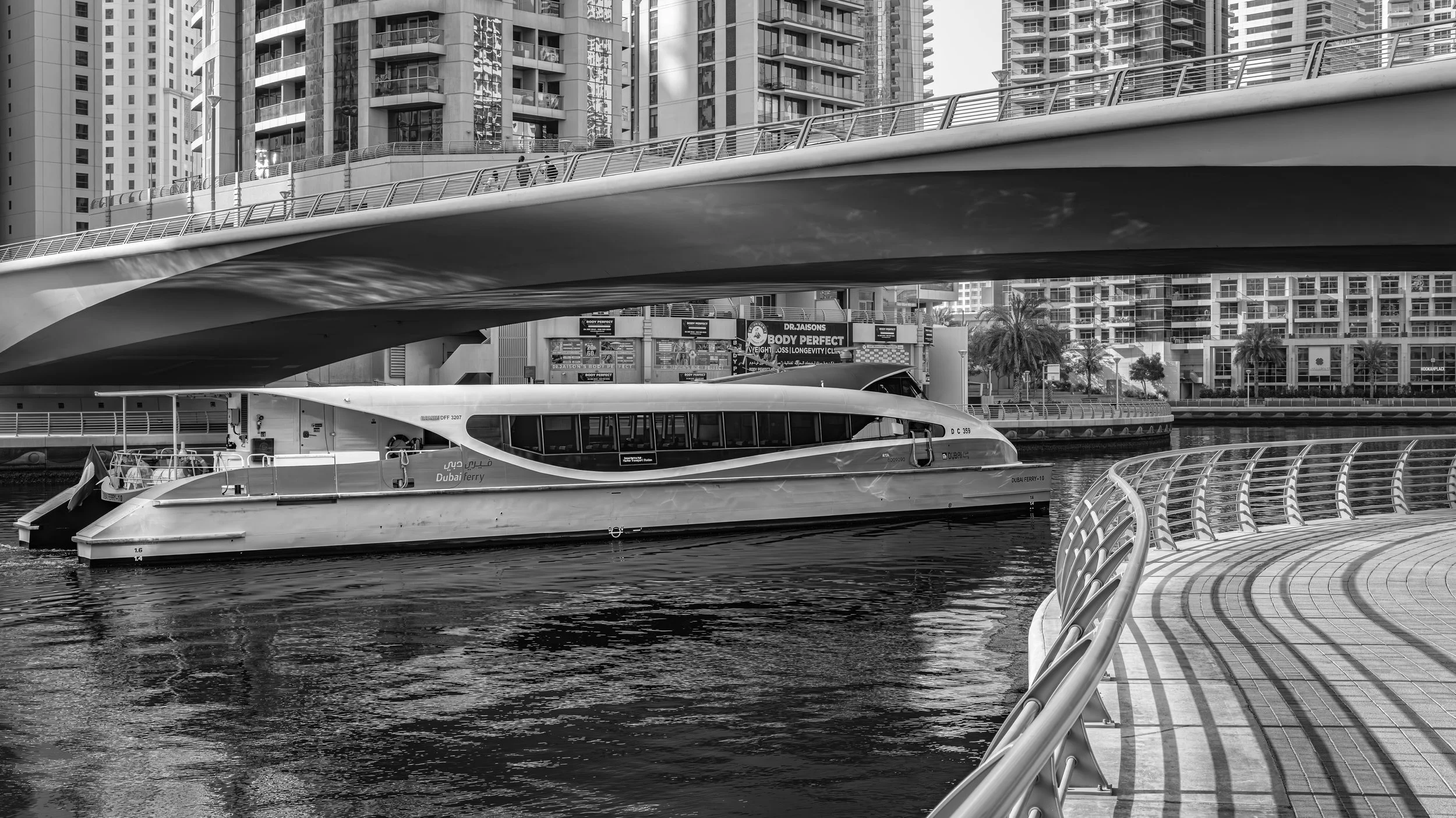 A cityscape with modern high-rise buildings, a waterway, and a Dubai ferry boat in the foreground, along with a curved walkway and railing along the water's edge. Dubai Marina, Dubai, UAE.