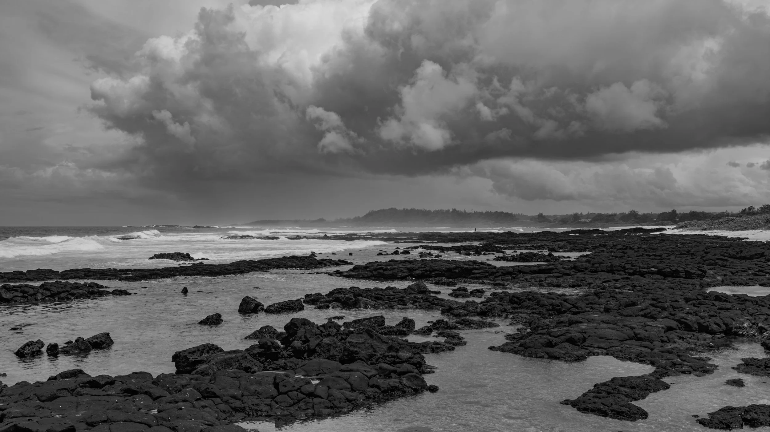 Black and white photograph of a rocky coastline with waves crashing against rocks, under a cloudy sky. In the distance, a person stands near the water's edge.