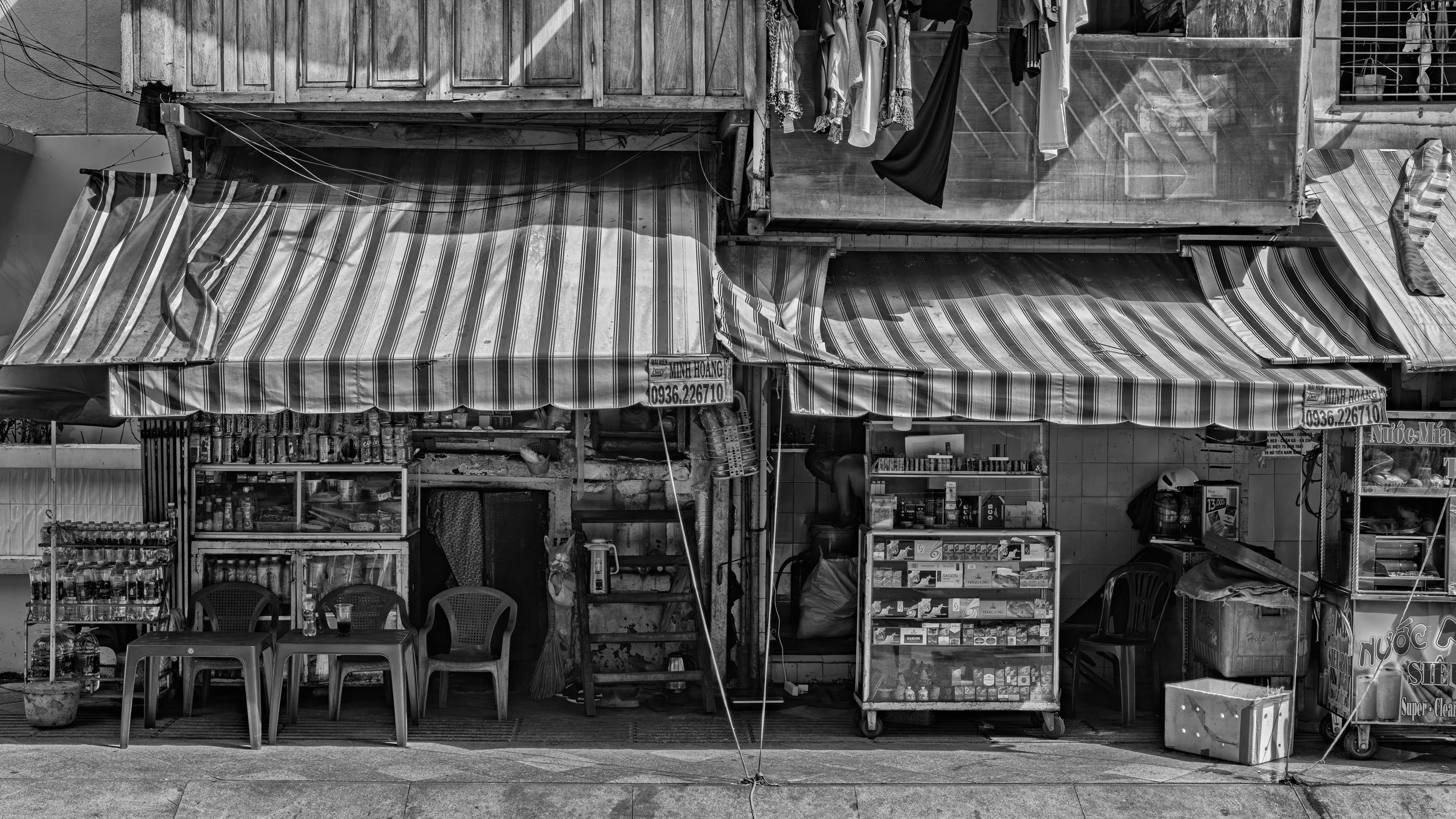 A black-and-white photo of a street-side small shop with striped awnings, outdoor chairs, and various displayed items including bottles and food boxes.