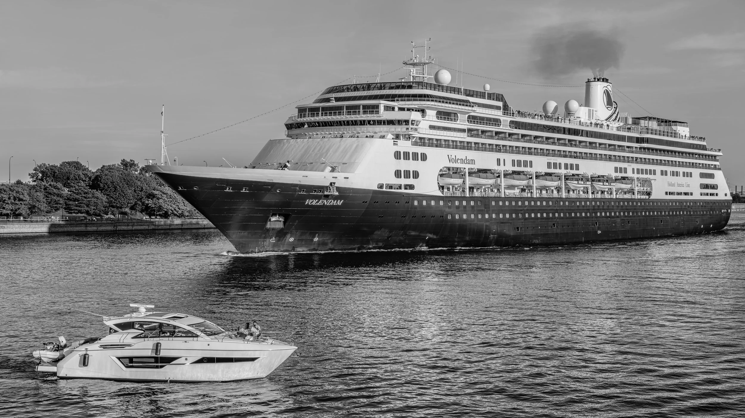 A large cruise ship named Volendam sailing on a body of water with smaller yachts in the foreground and trees lining the shore in the background.
