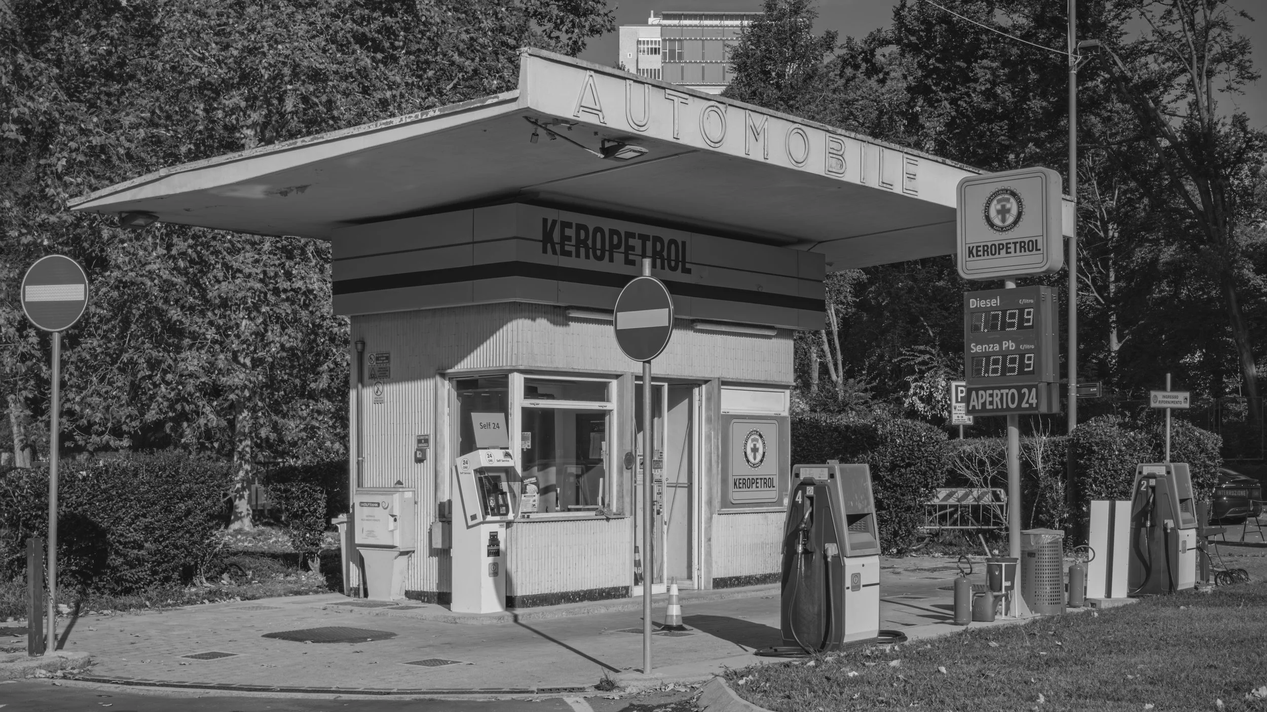 A black and white photo of a small fuel station with multiple gas pumps, surrounded by trees and some buildings in the background. The station has a sign indicating diesel fuel price and an electronic board with prices for Diesel and gasoline. The st