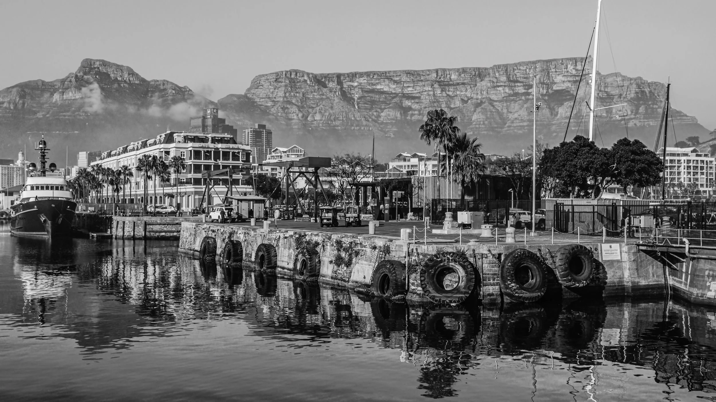 Black and white photo of a harbor with a boat docked, industrial buildings, trees, and a mountainous backdrop. Cape Town, South Africa.