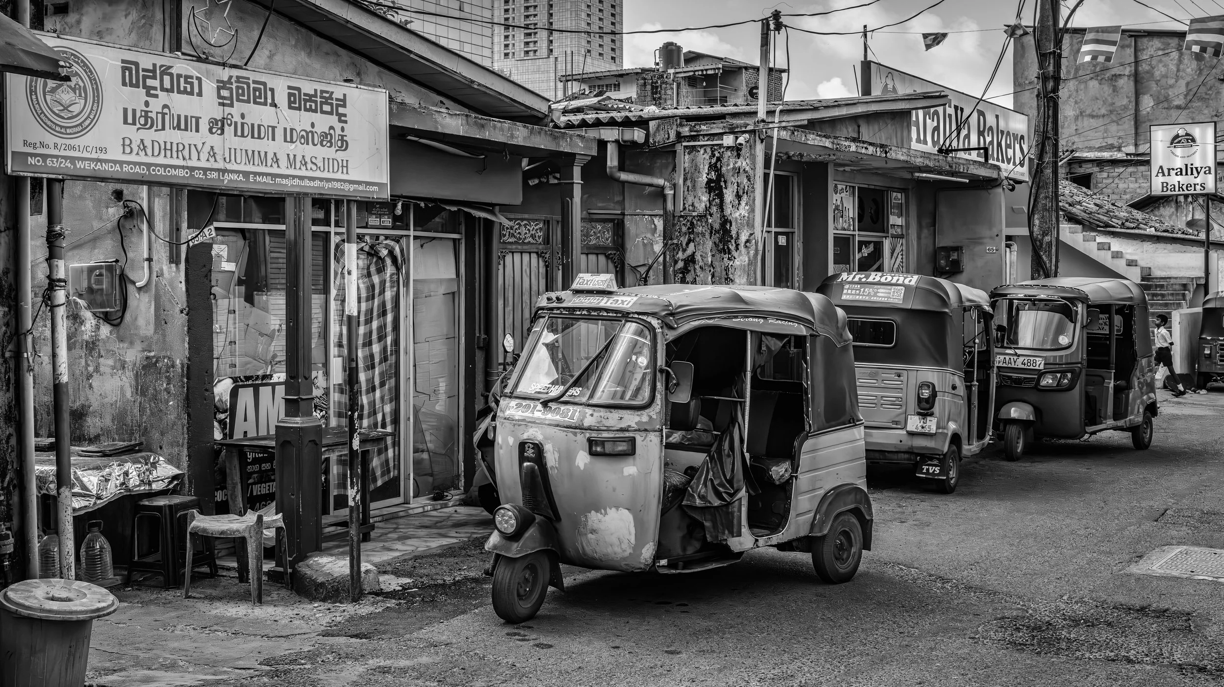 Street scene in an urban area with parked auto-rickshaws, small shops, and signs, including a sign for 'Araliya Bakers.' A man is walking in the background, and high-rise buildings are visible in the distance. The image is in black and white.