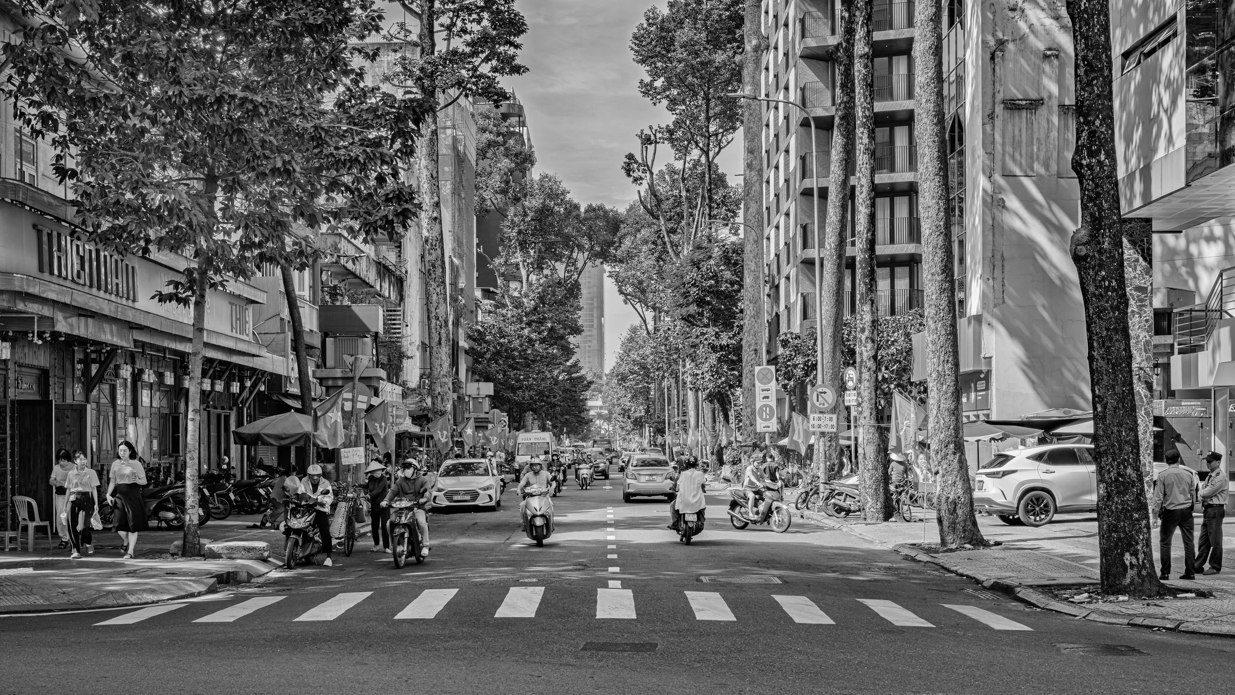 Black and white city street scene with cars, motorcycles, and pedestrians. Tall buildings line both sides, with trees along the sidewalk. People walk, some wearing masks, and market stalls with umbrellas are visible on the left side.