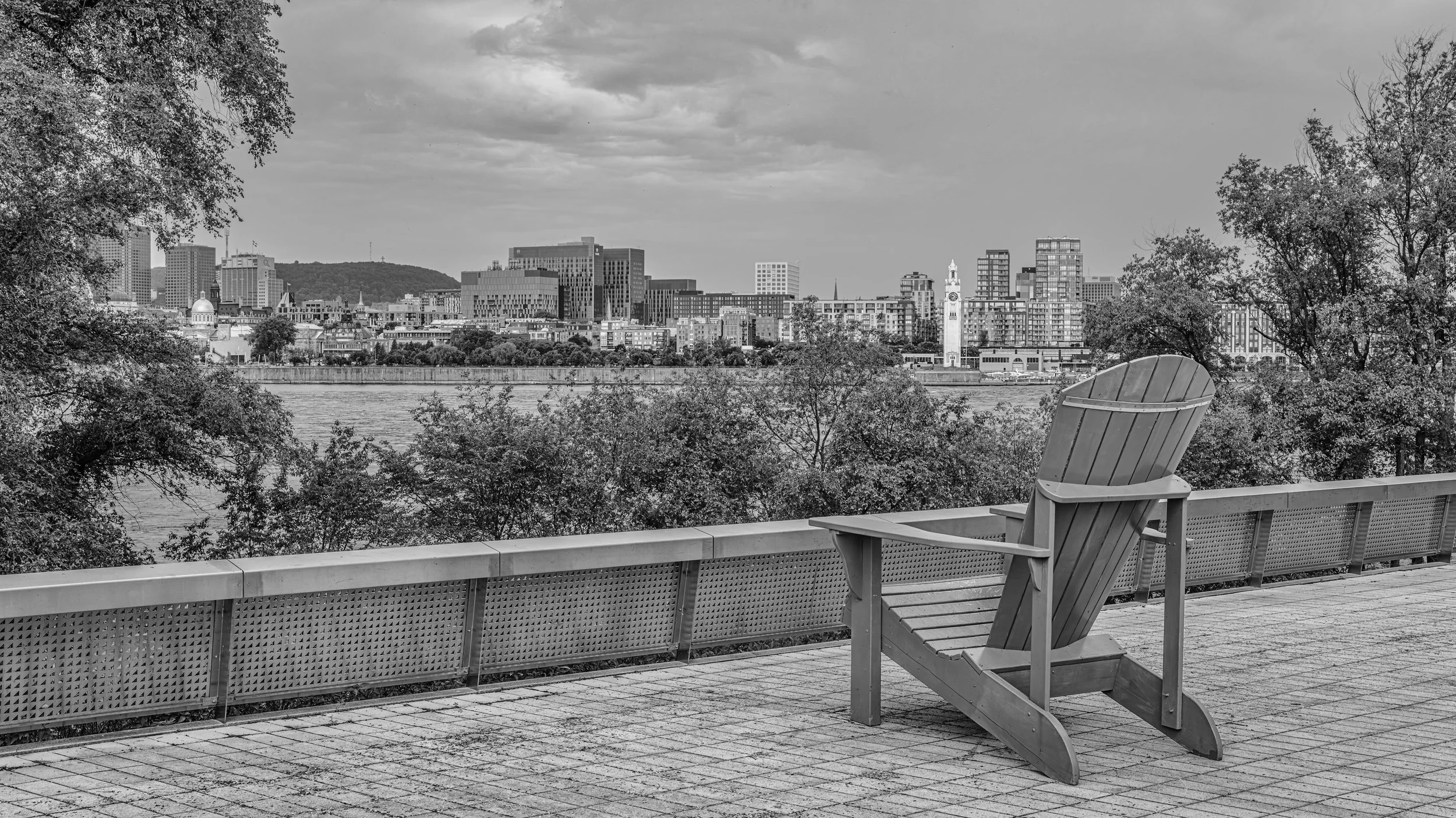 A black and white photo of a city skyline viewed from a waterfront park. There is a wooden chair on a paved area in the foreground, trees on either side, and a large river or body of water separating the park from the city. The city buildings include