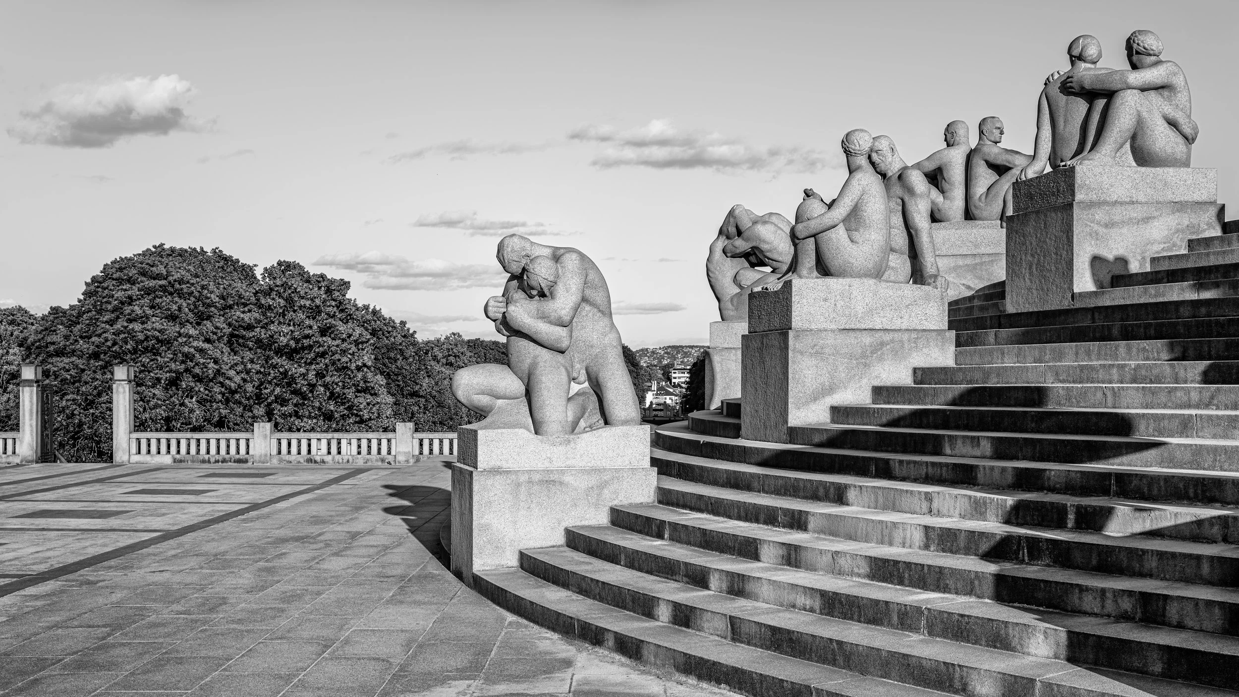 A series of stone sculptures depicting human figures in various postures, positioned on a raised platform with stairs. Trees and a cloudy sky are in the background.