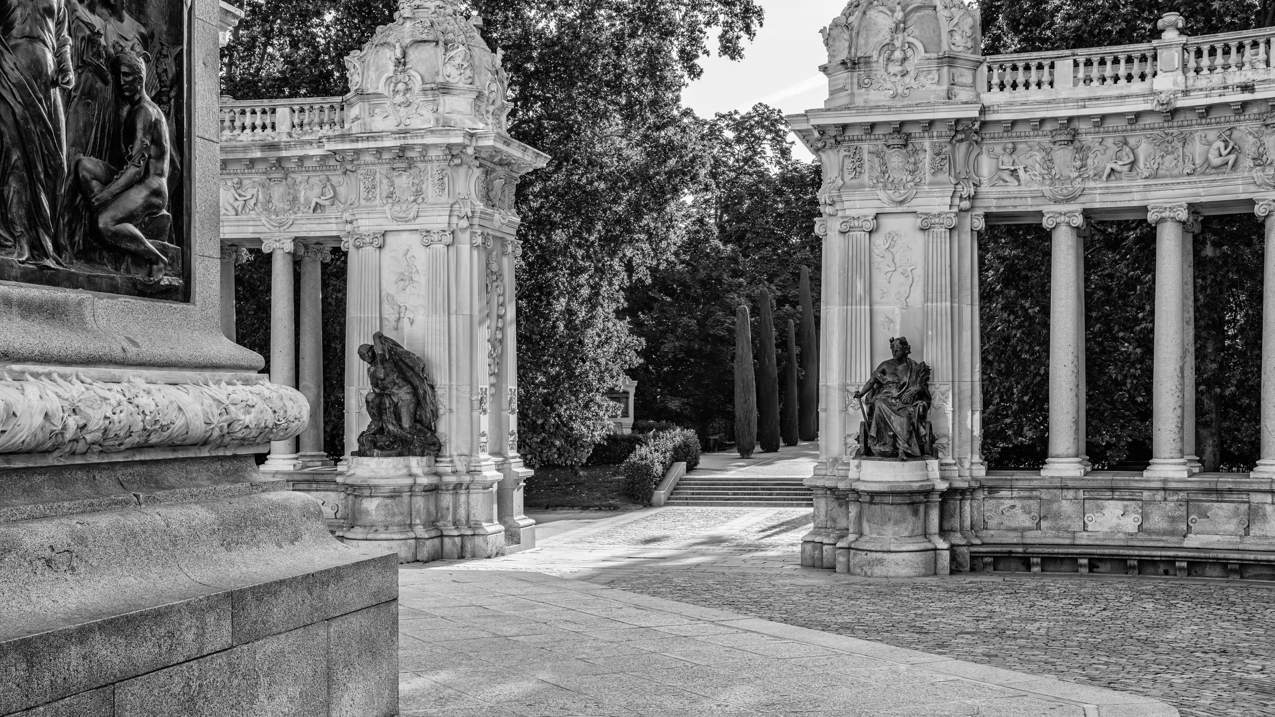 Black and white photo of an ornate stone archway with sculptures of seated women and floral decorations. Trees and foliage in the background.