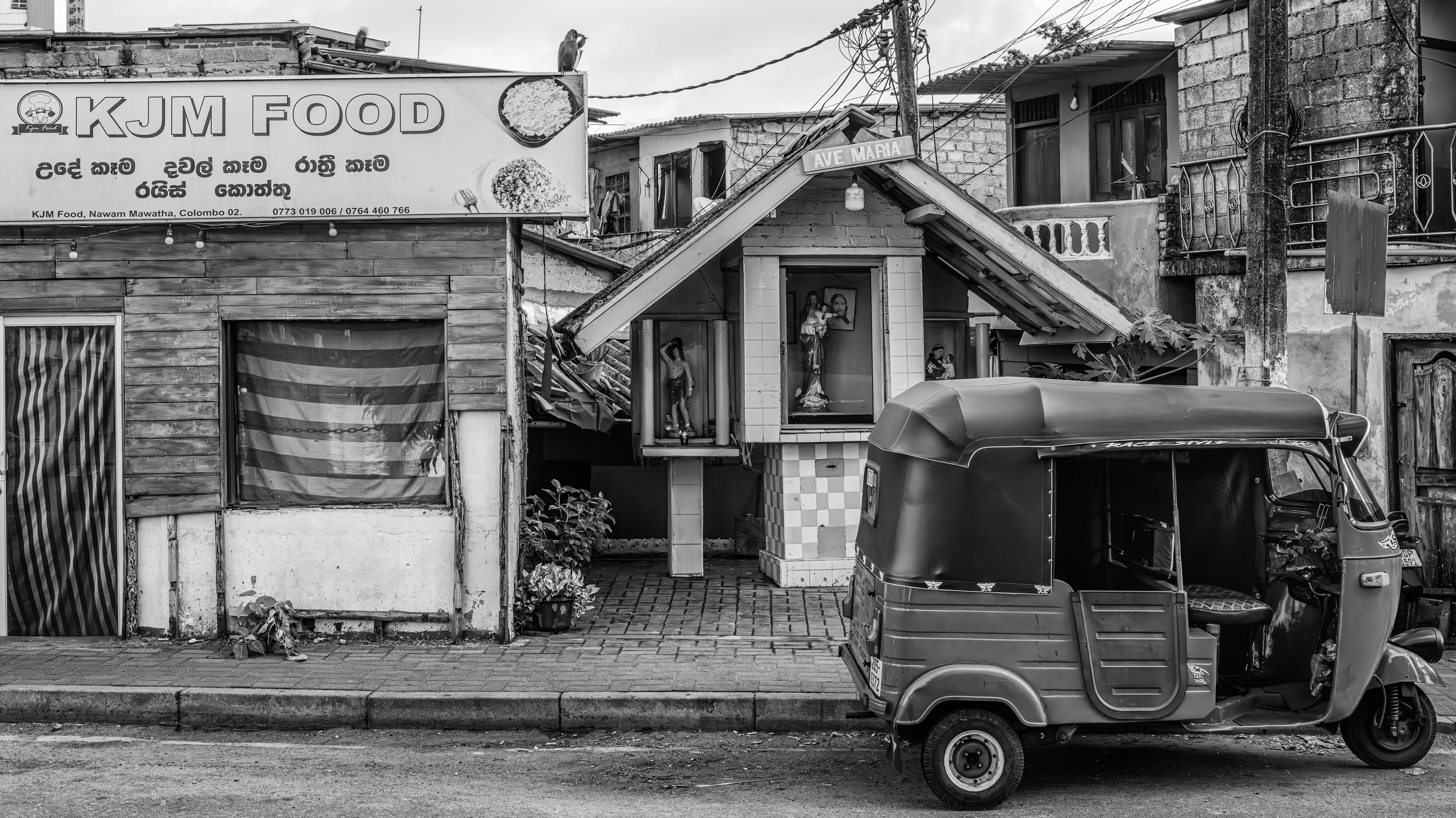 A black and white photo of a street scene featuring a small building with religious statues and a vehicle parked in front. There is a sign for KJM Food and a street sign for Ave Maria.