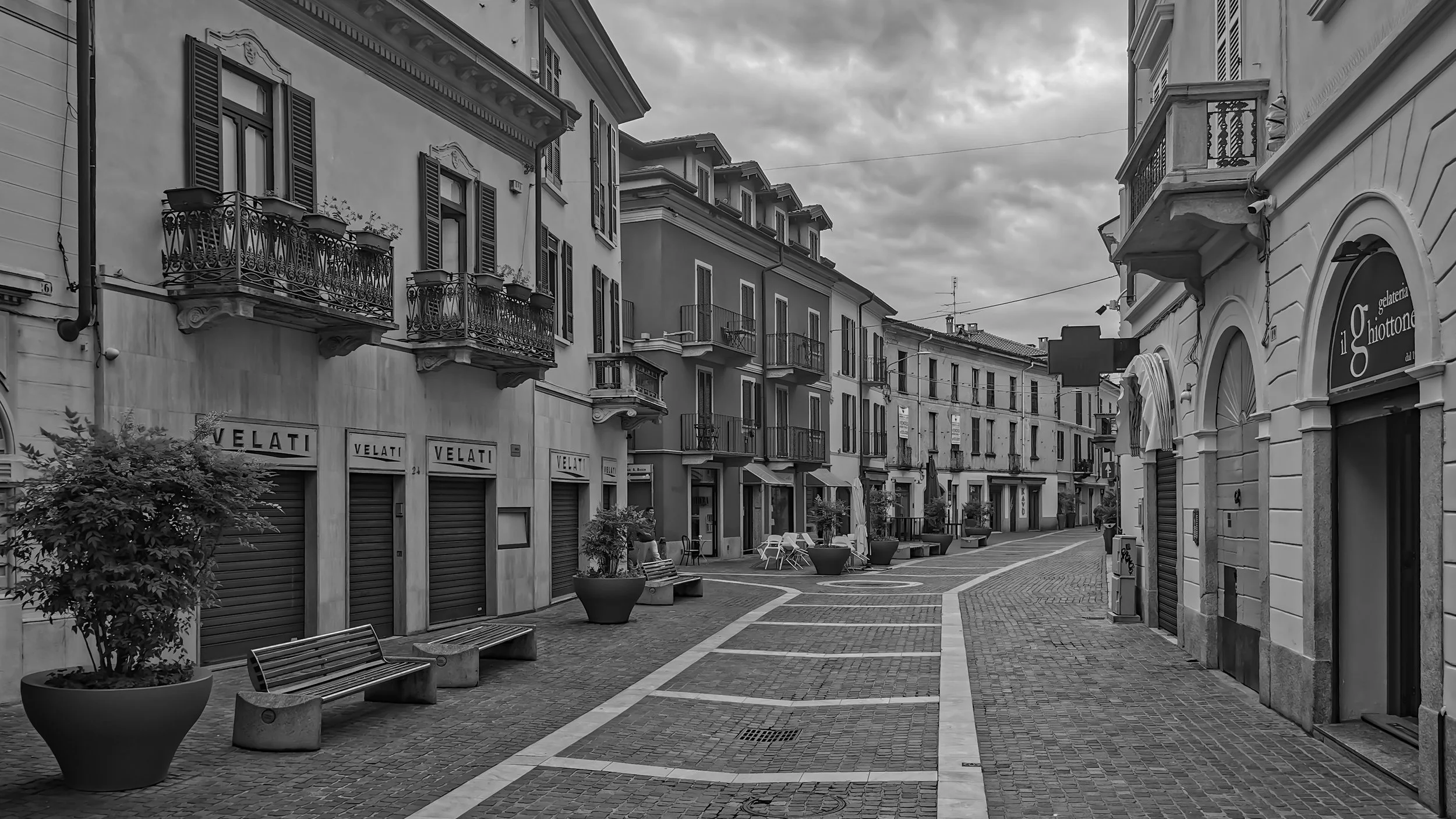 Empty European-style street with benches, potted plants, and closed shops, under an overcast sky.